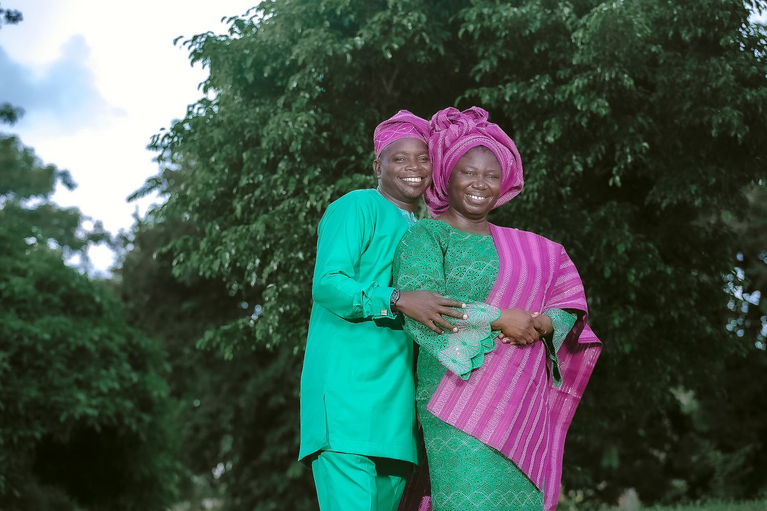 A couple in traditional African attire embraces outdoors, wearing matching green and pink garments.