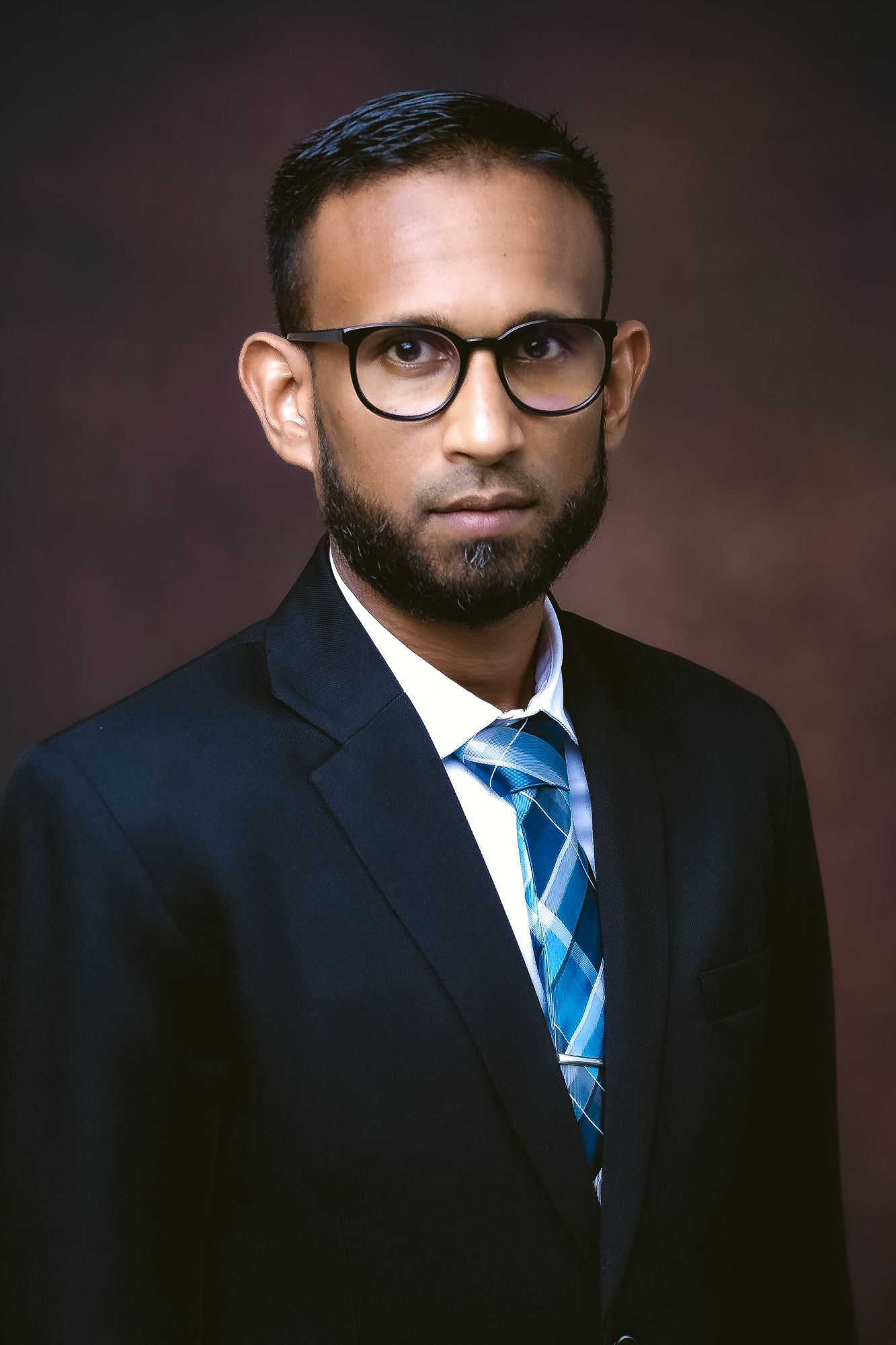 Professional headshot of person in dark suit with blue striped tie and glasses against dark background.