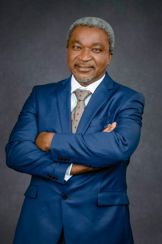 Professional portrait of a person in a navy blue suit with a patterned tie standing with arms crossed against a dark backdrop.