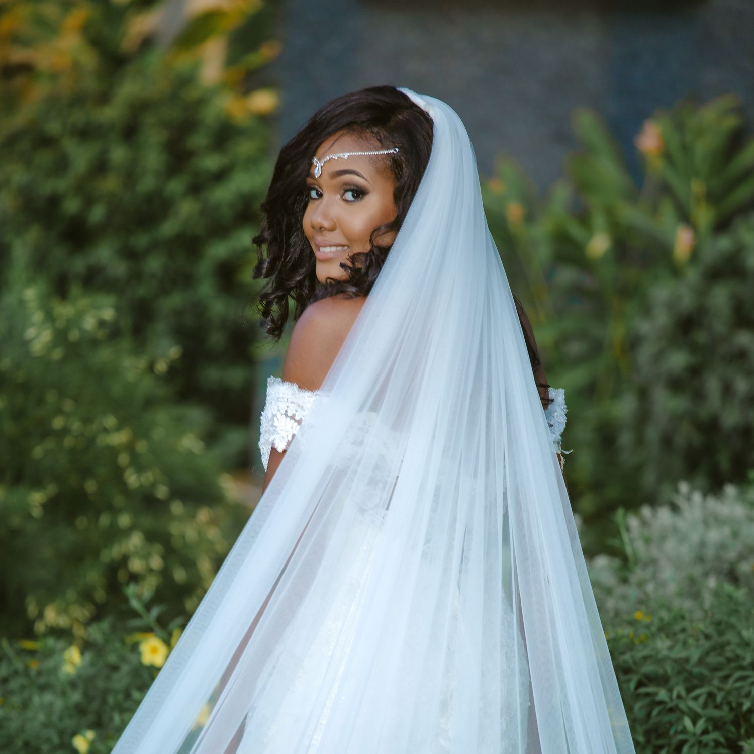 A beautiful bride in a white strapless gown and flowing veil poses elegantly in a garden setting.