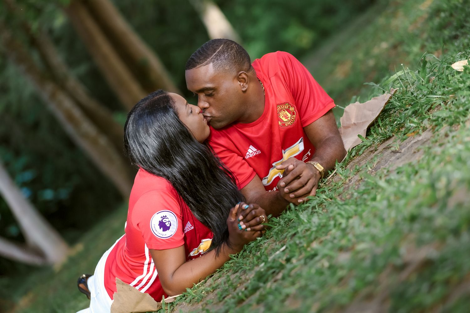 Couple in matching red Manchester United jerseys lying together on grassy hill slope.