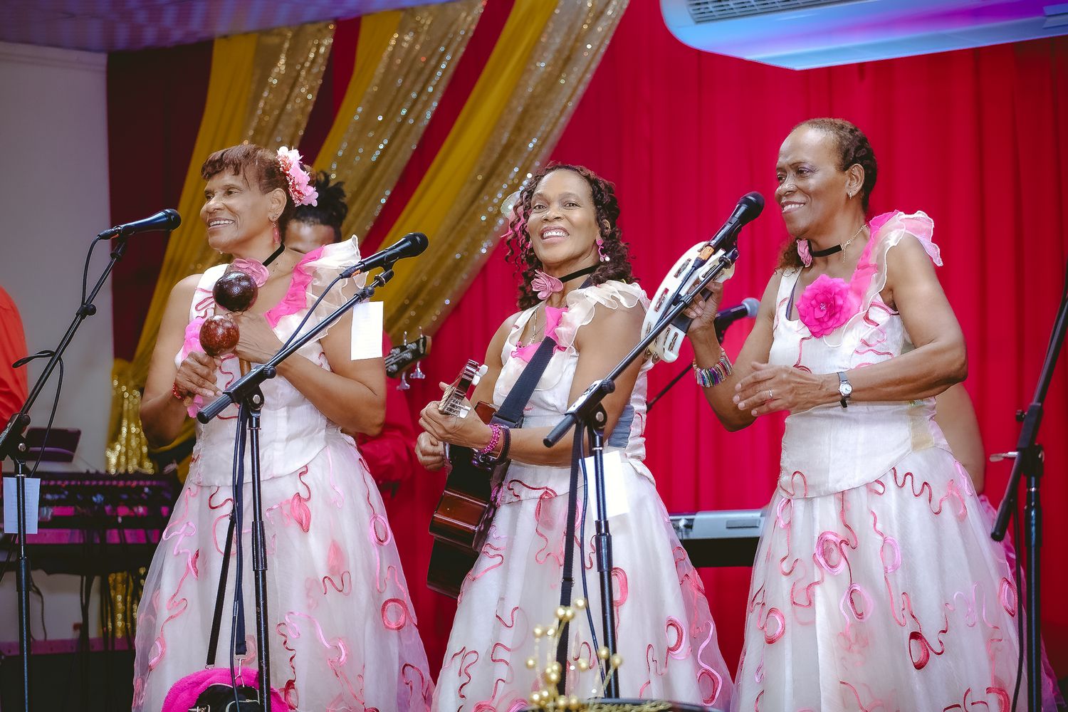 Three performers in white floral dresses sing together on stage with microphones at a musical event.