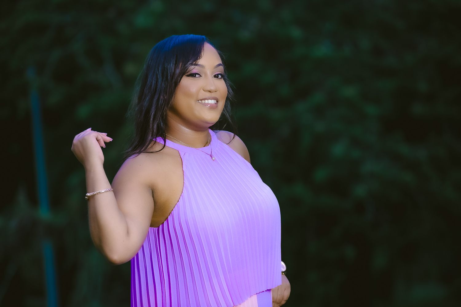 Woman in purple pleated halter dress poses against dark foliage.