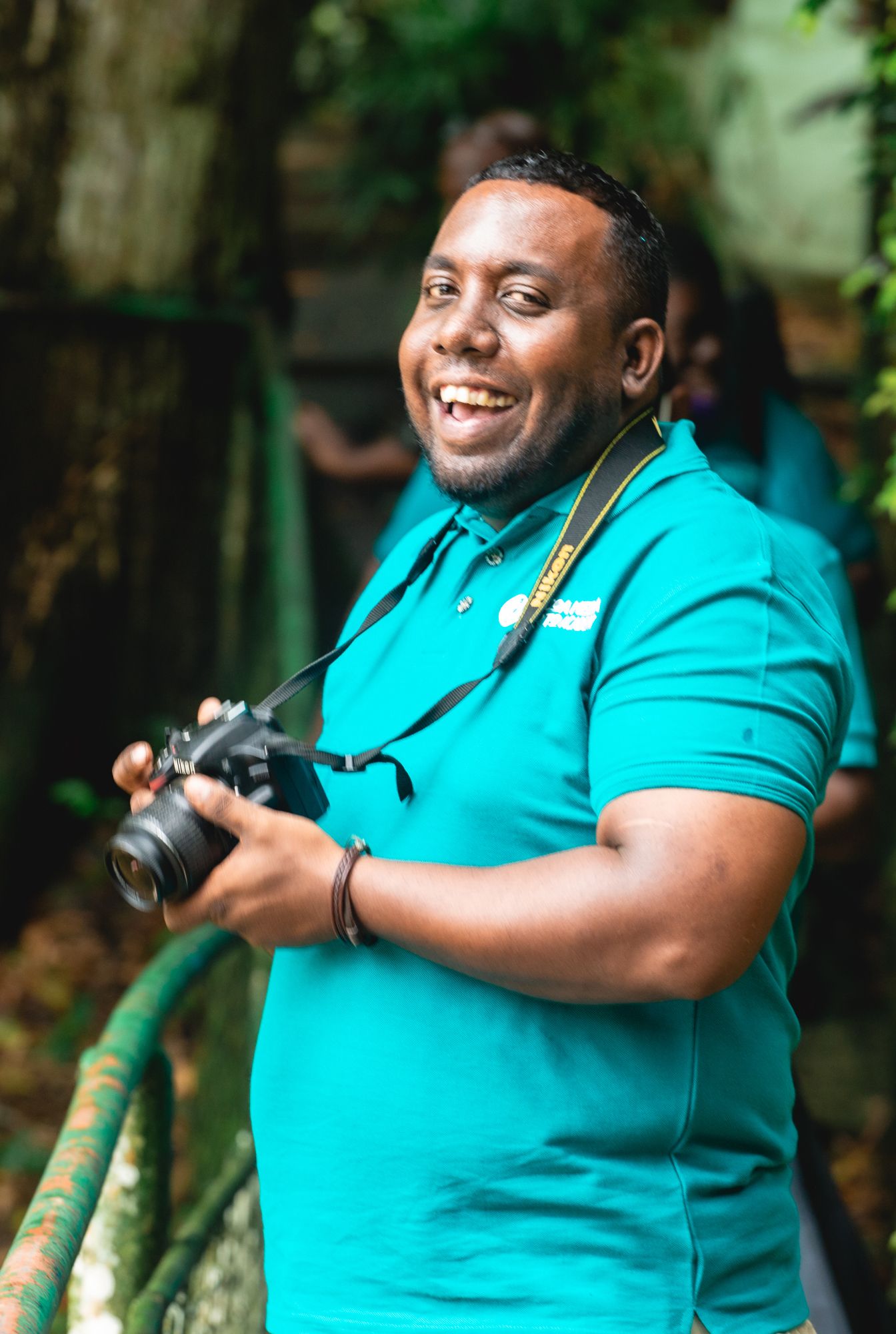 A photographer in a bright turquoise polo shirt smiles while holding a camera outdoors in a natural setting.