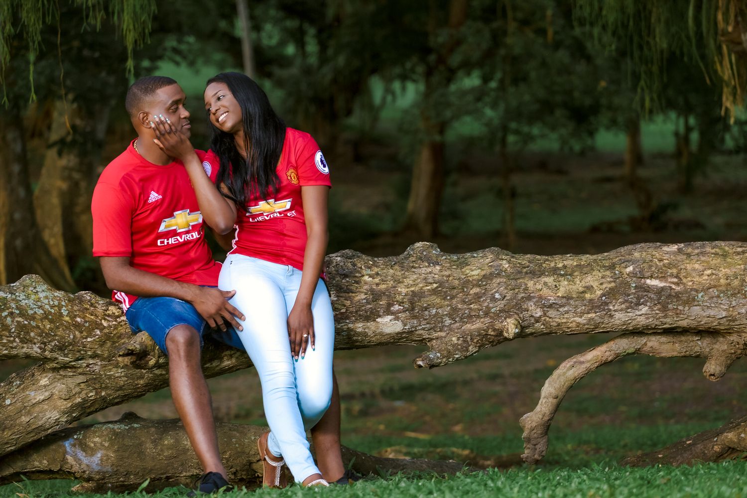 A couple wearing matching red shirts share a moment while sitting on a fallen tree in a park.