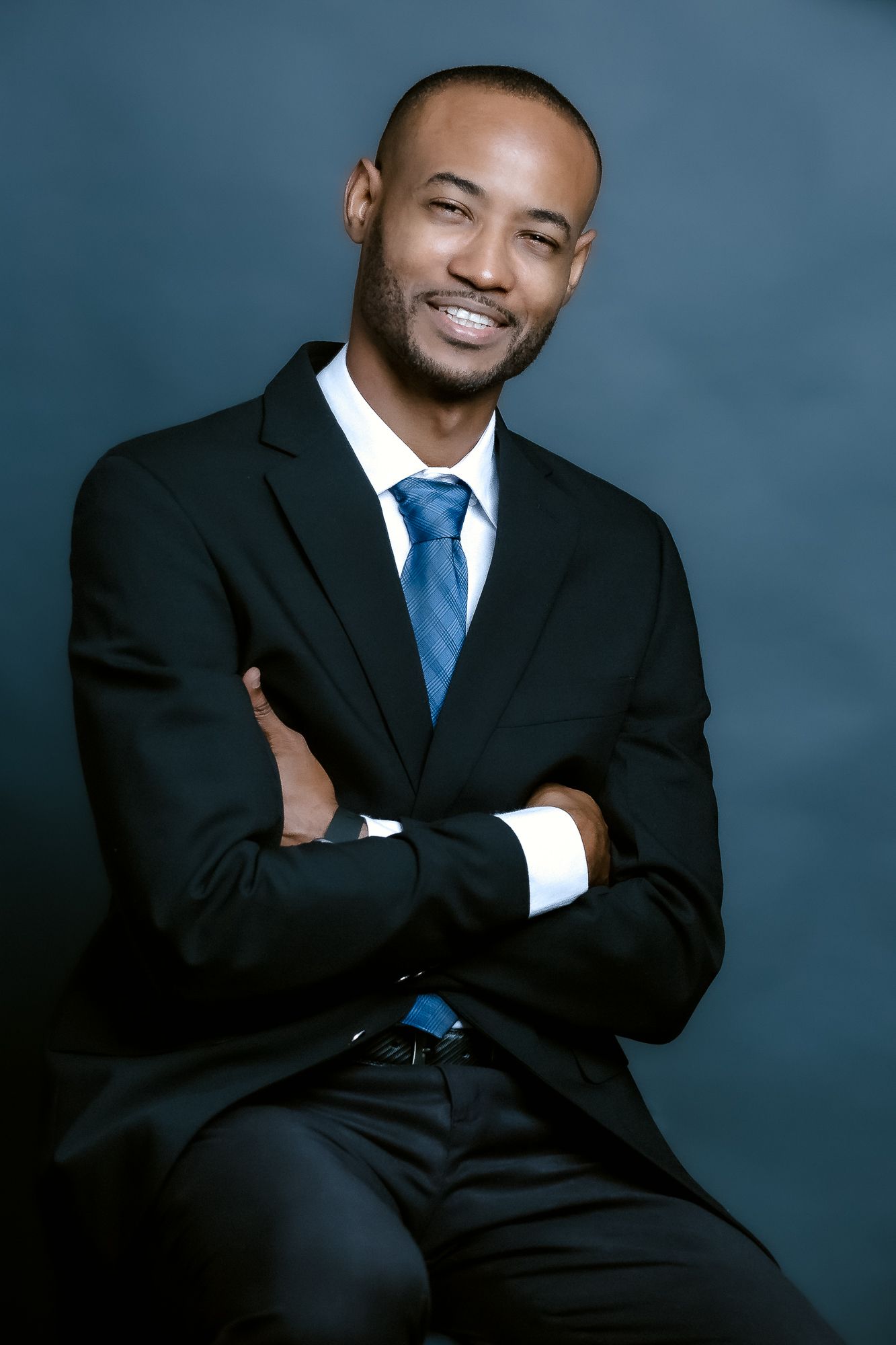 Professional portrait of person in black suit with blue tie seated against blue background.