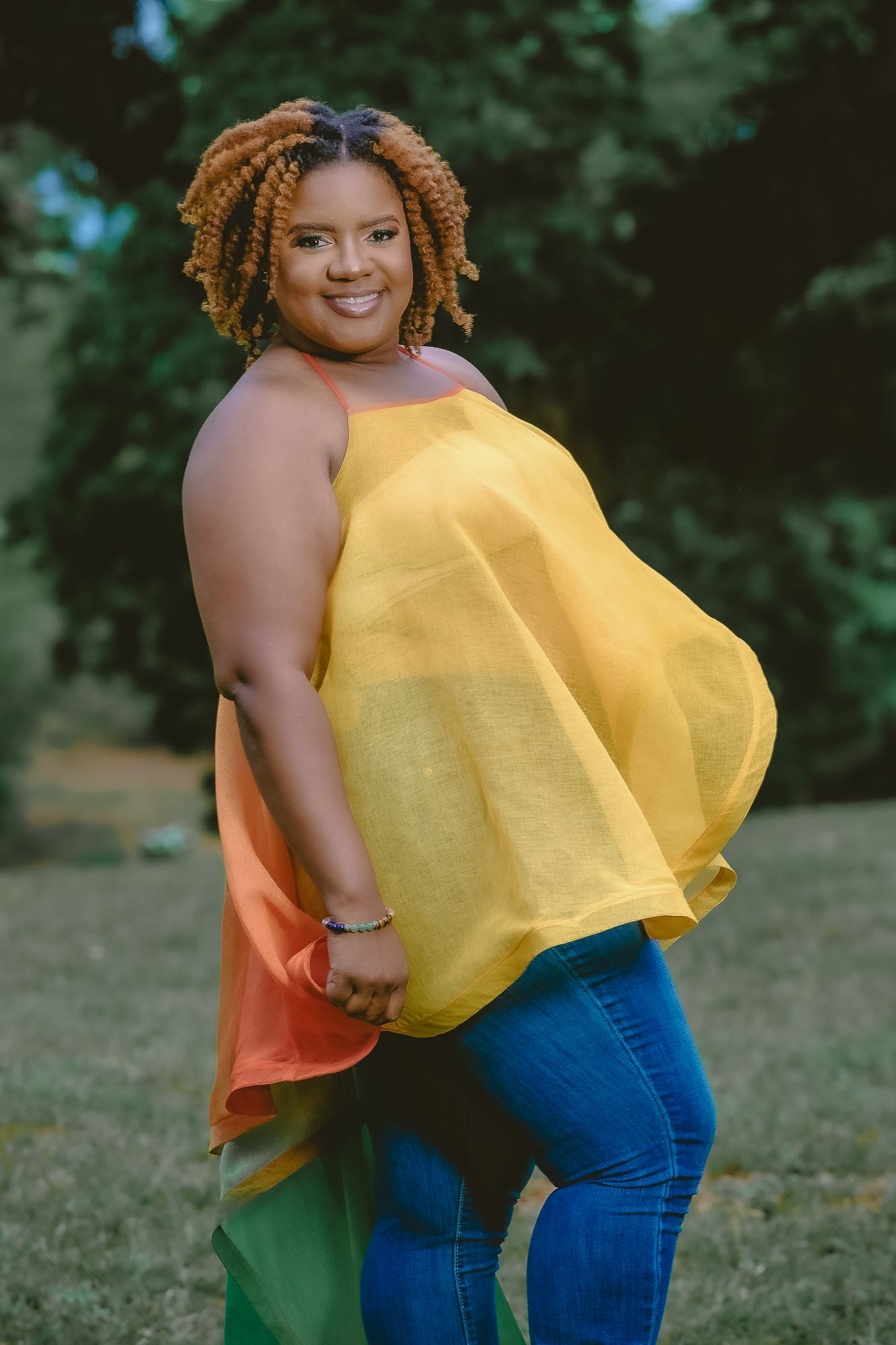 Person in flowing yellow top and jeans poses against green outdoor background.
