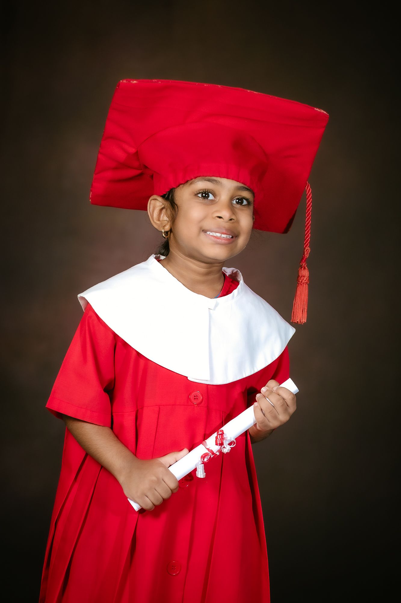 Young graduate in red cap and gown holds diploma while smiling at camera during graduation photos.