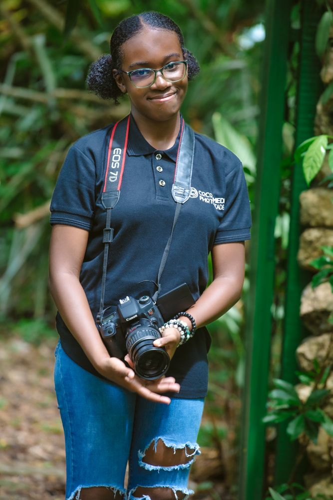 A photographer with a DSLR camera poses in front of bamboo trees while wearing casual attire.
