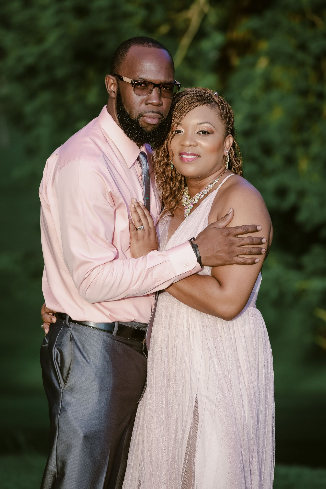 A couple in coordinating pink outfits pose together against a lush green background.