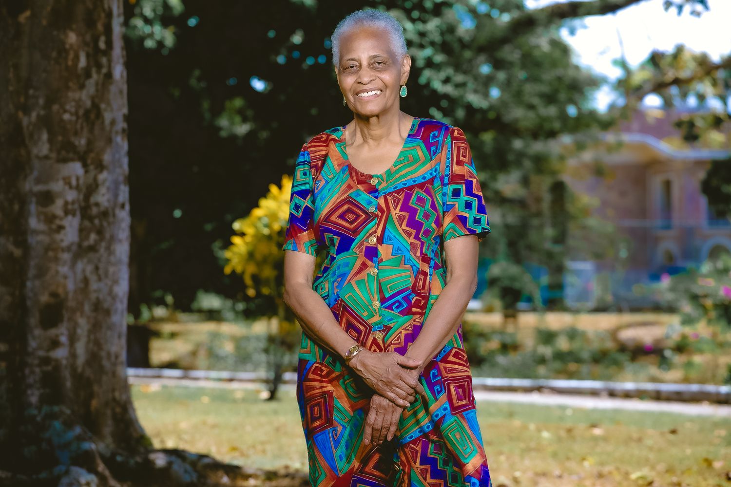 Portrait in vibrant geometric print dress standing under large tree in park setting.