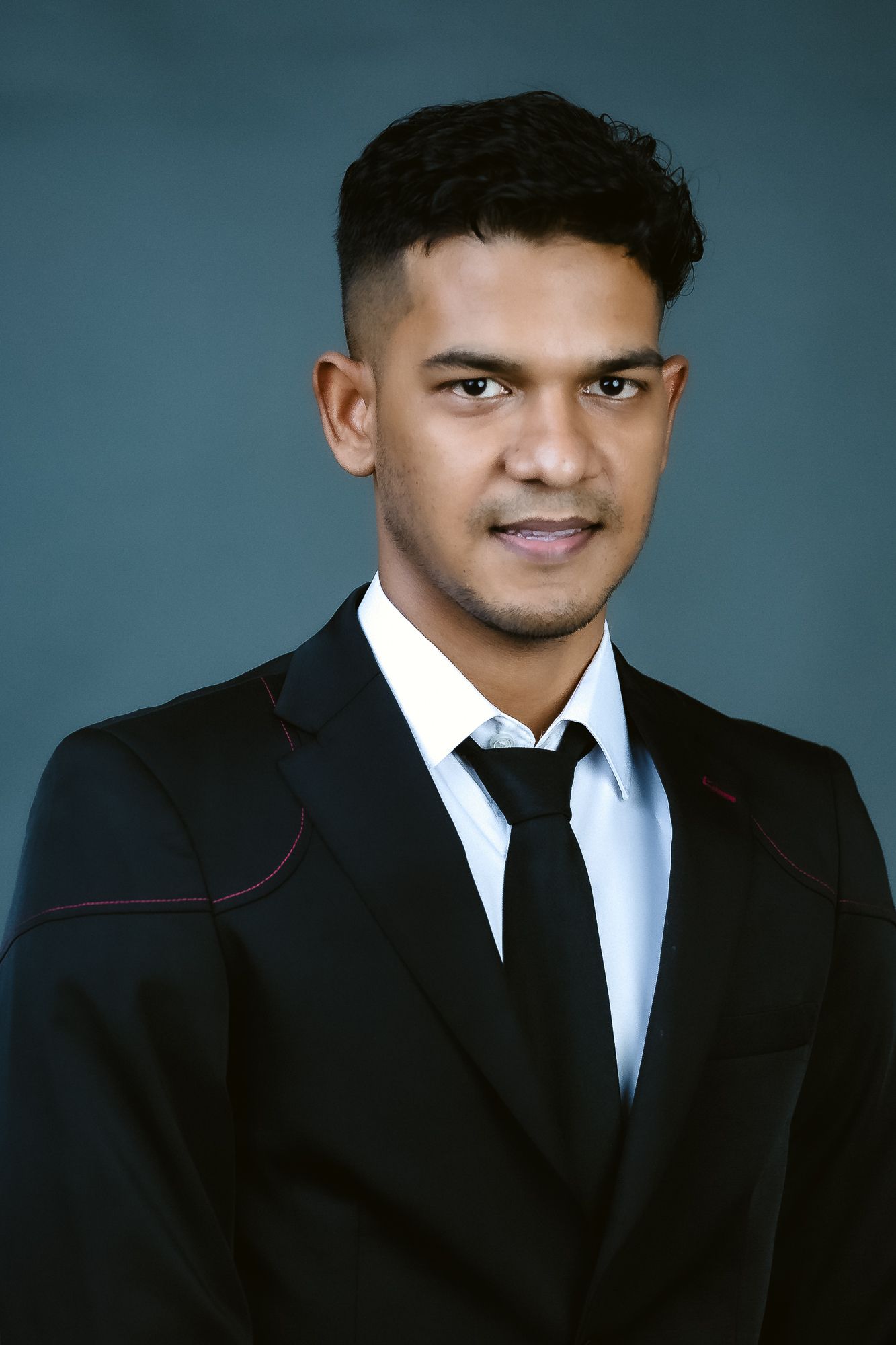 Studio headshot of a person wearing a black suit and tie against a blue backdrop.