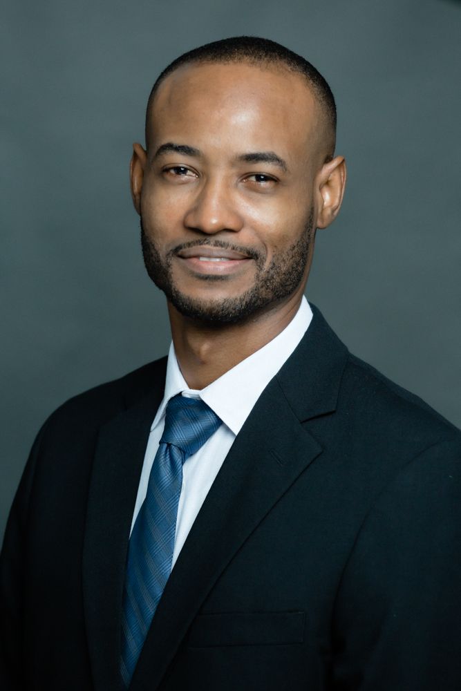 Professional headshot portrait of a smiling person in a dark suit and blue tie against a teal background.