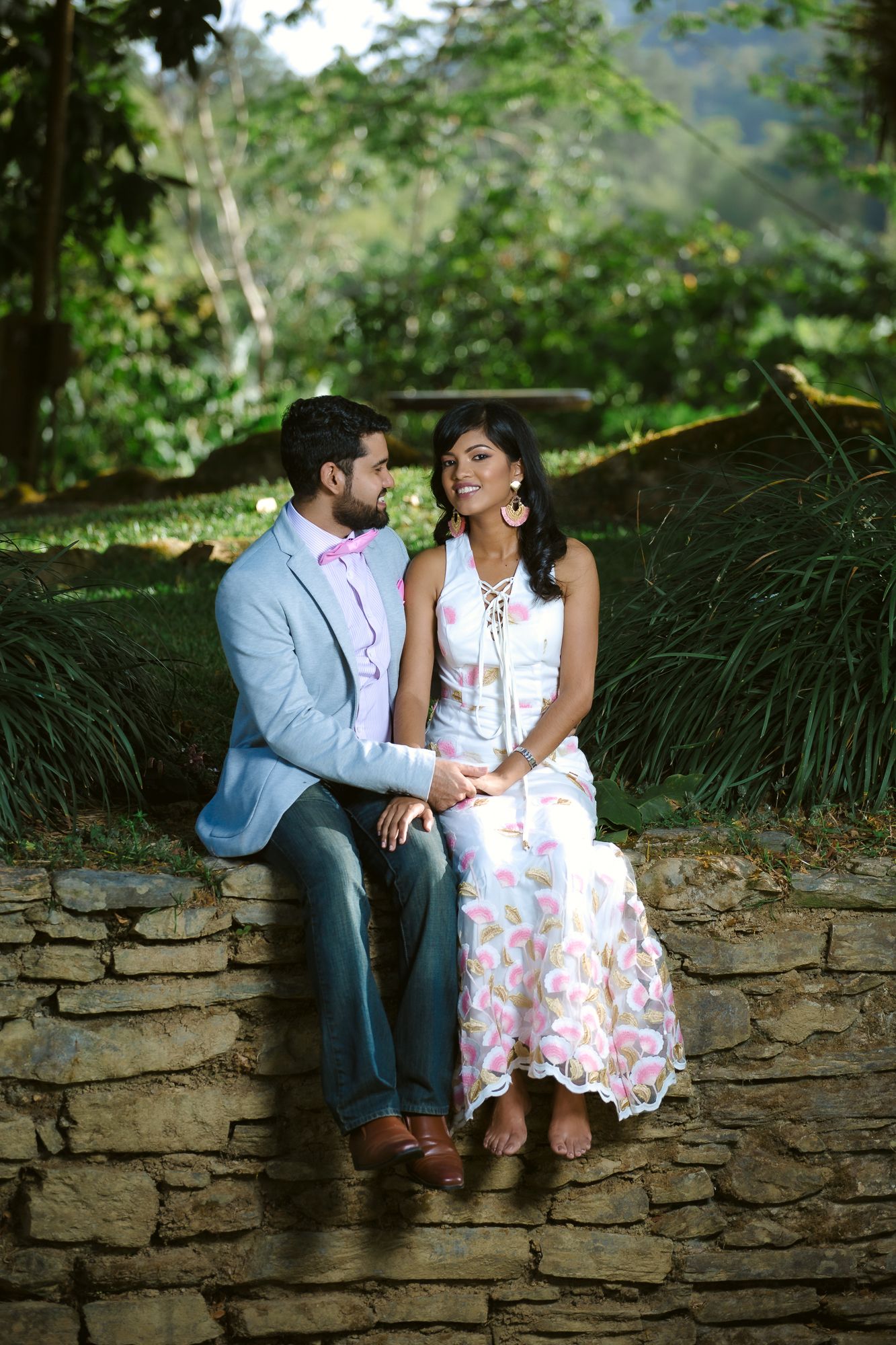 A couple in wedding attire sits together on a rustic stone wall surrounded by lush greenery on a sunny day.