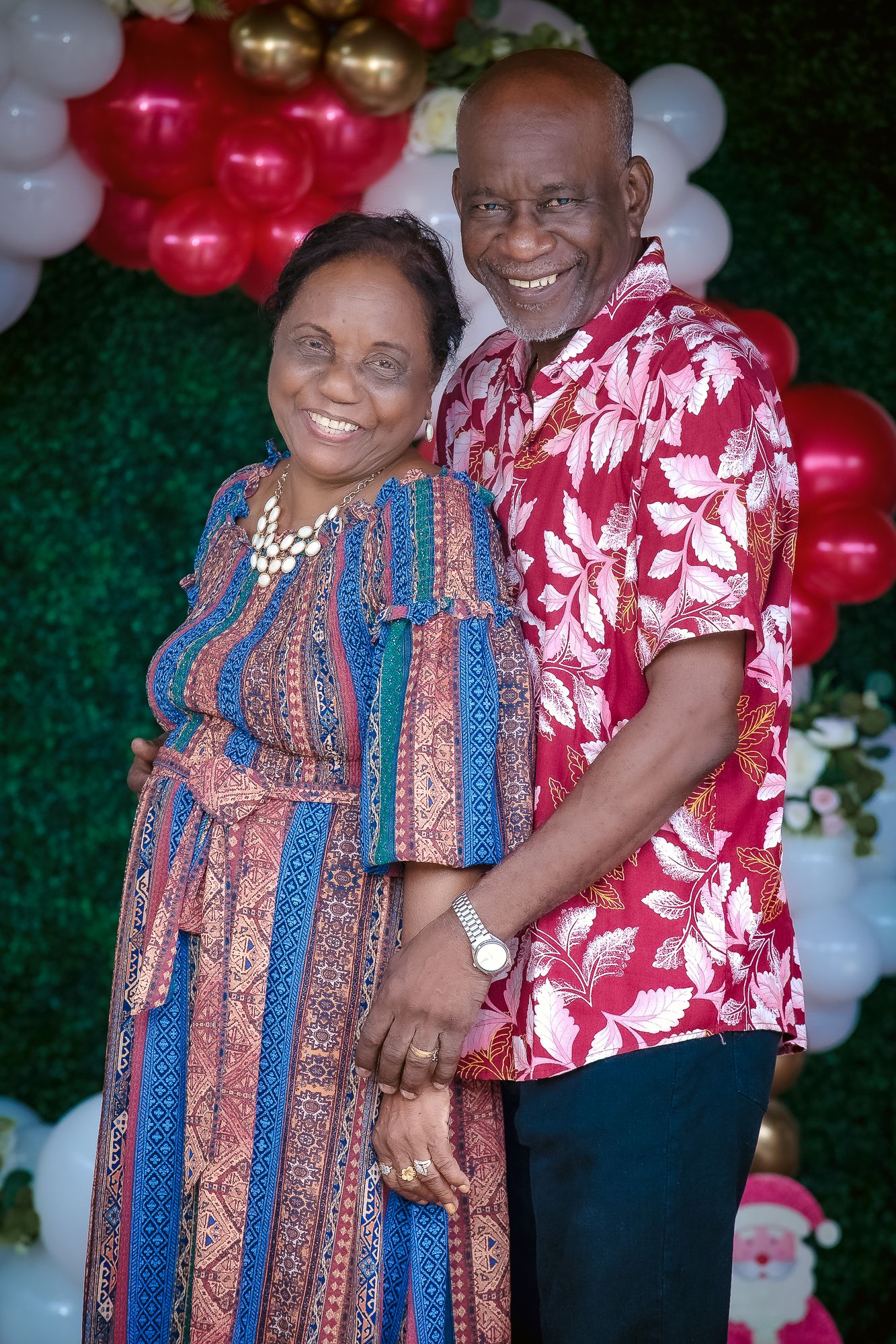 A couple in vibrant patterned clothing poses together at a celebration with balloon decorations.