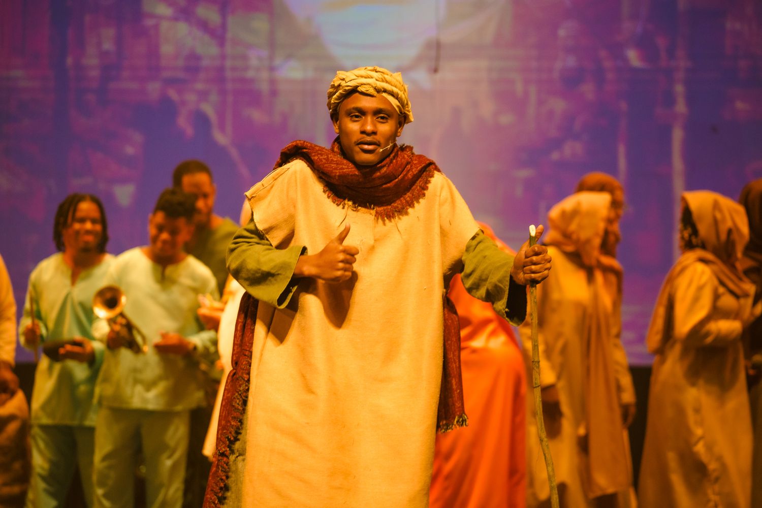 Performers in colorful robes singing on stage during a theatrical production with dramatic lighting.