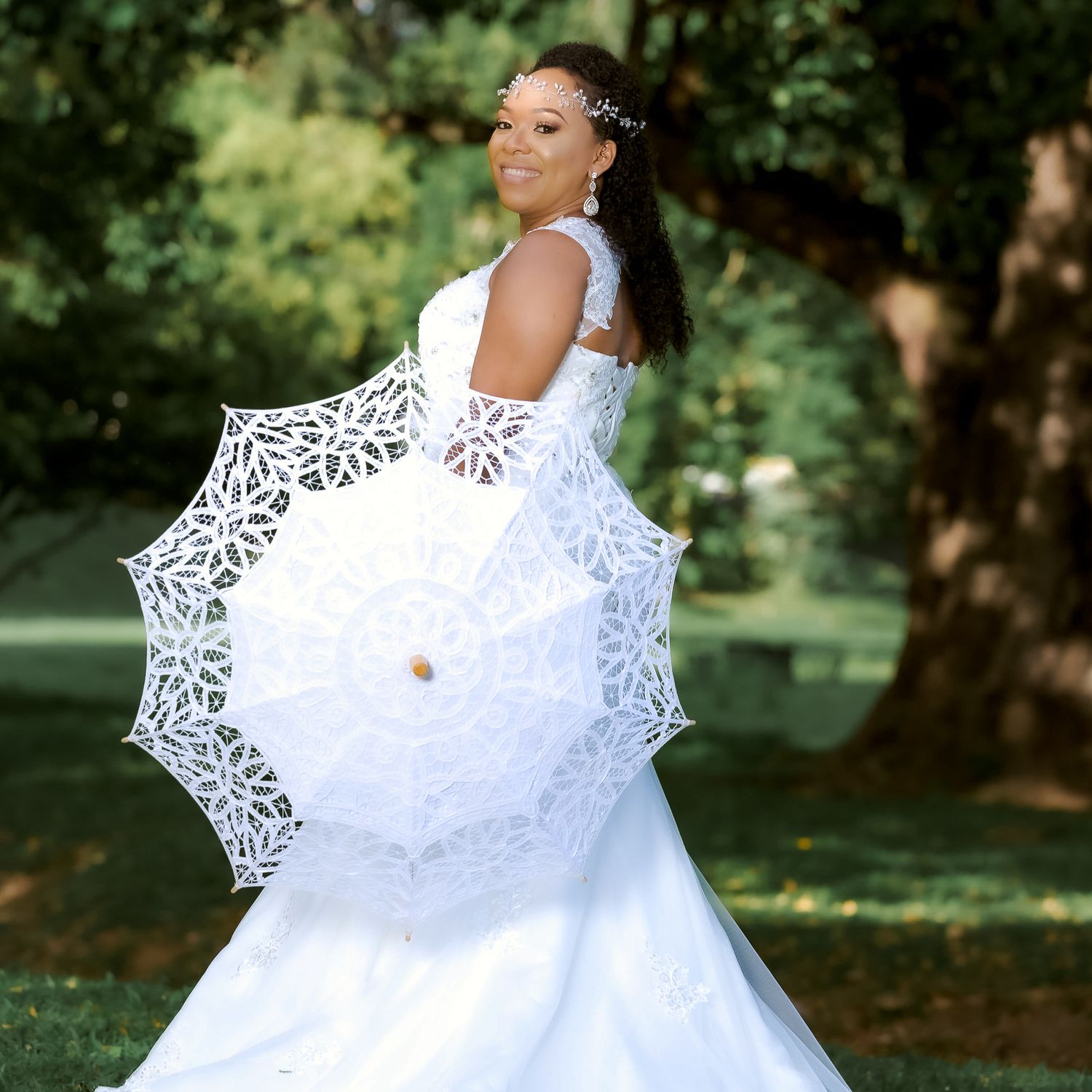 A bride holds a delicate white lace parasol while posing in her fitted wedding gown outdoors.