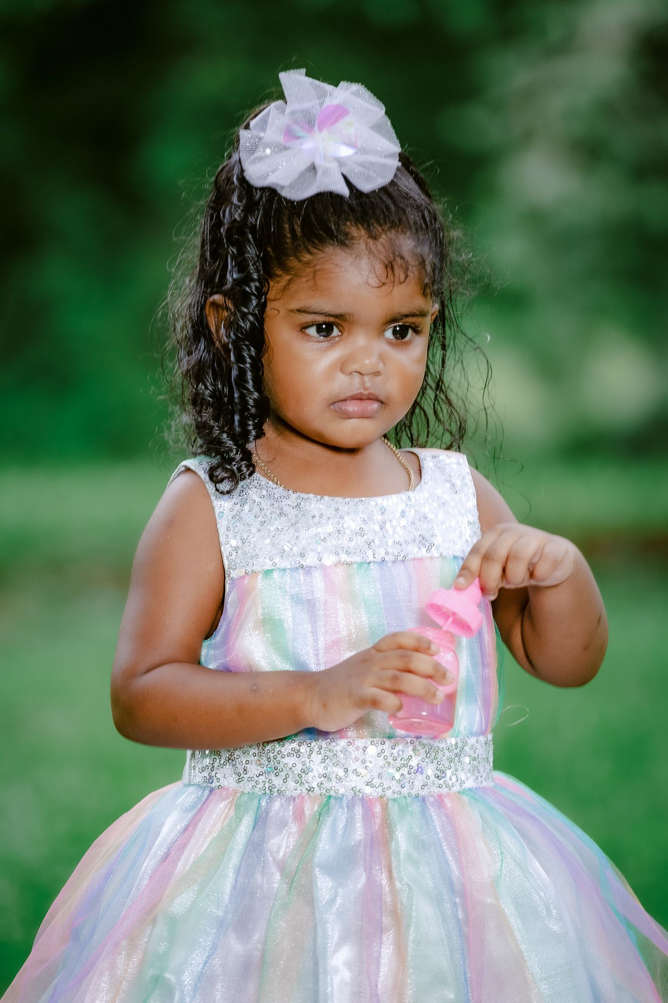 A young girl in a sparkly pastel dress with a white flower hair accessory poses in a garden.
