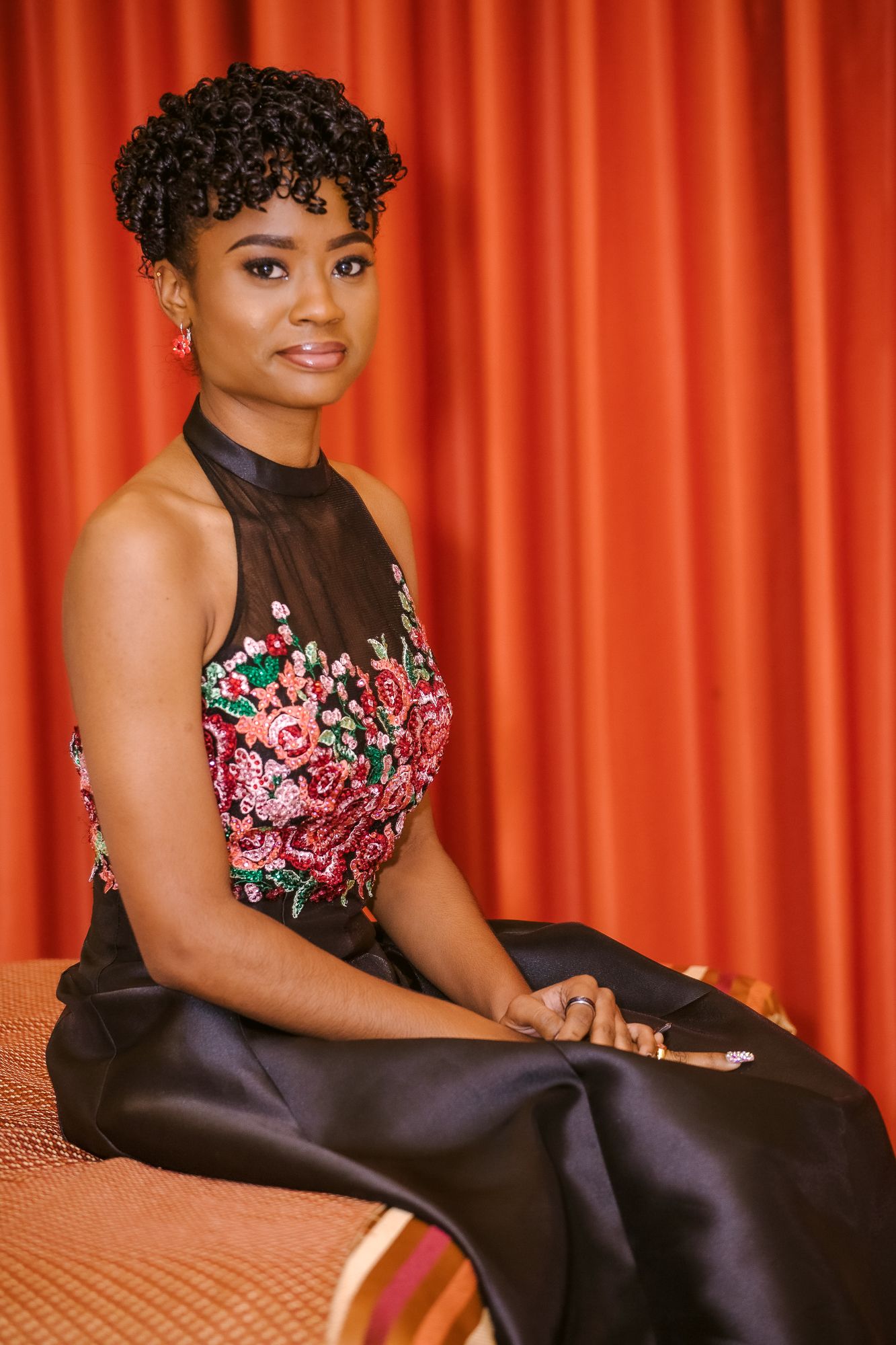 Portrait against orange curtain background wearing elegant black dress with floral embroidery.