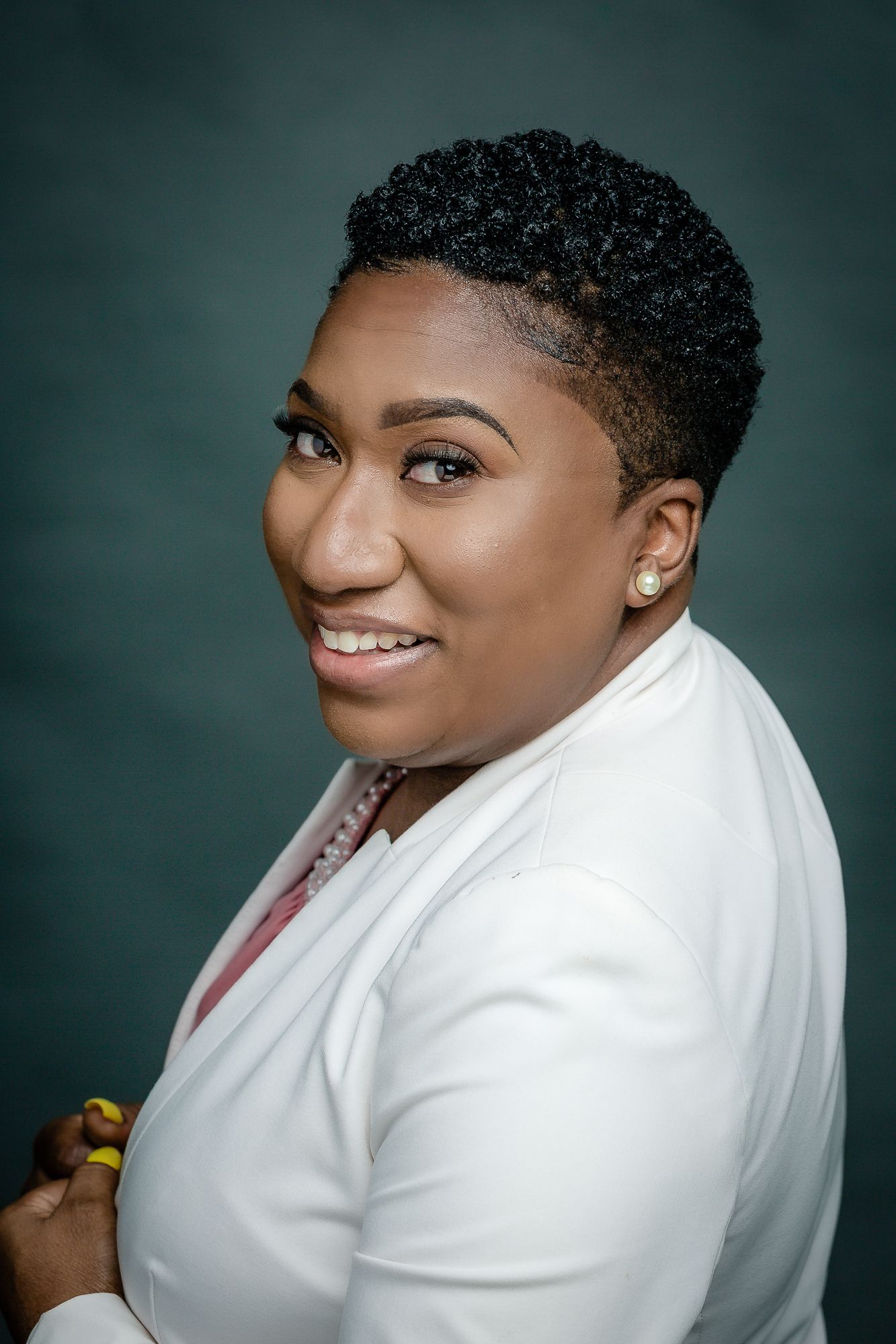 Professional studio portrait of a smiling person in a white blazer against a dark teal background.