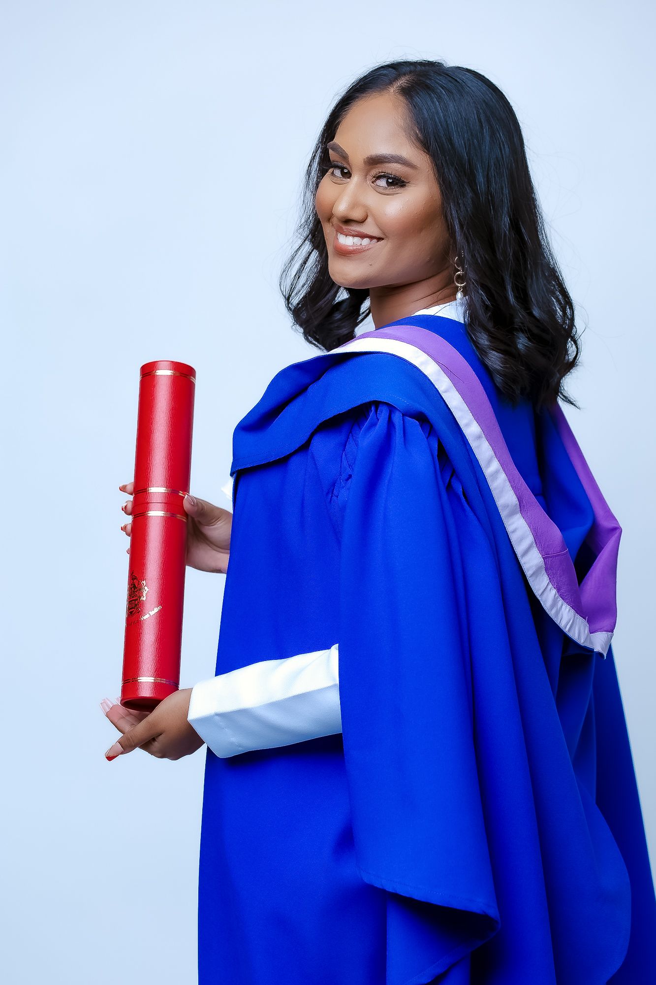 Graduate in blue and white graduation robes holding red diploma case with joyful expression.