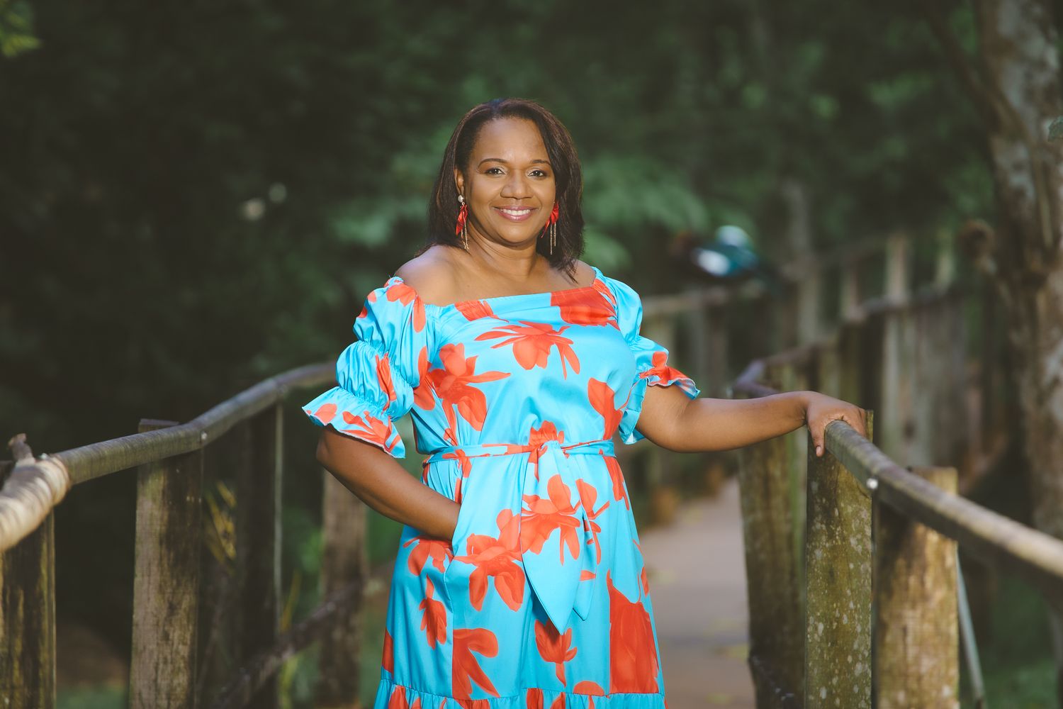 Woman in vibrant turquoise and orange floral dress poses on wooden bridge in nature setting.