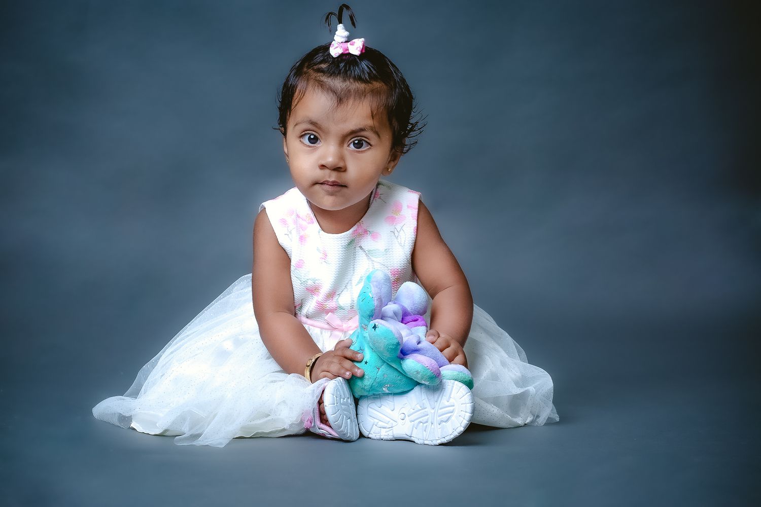 A small child in a white dress sits on gray backdrop holding a stuffed toy.