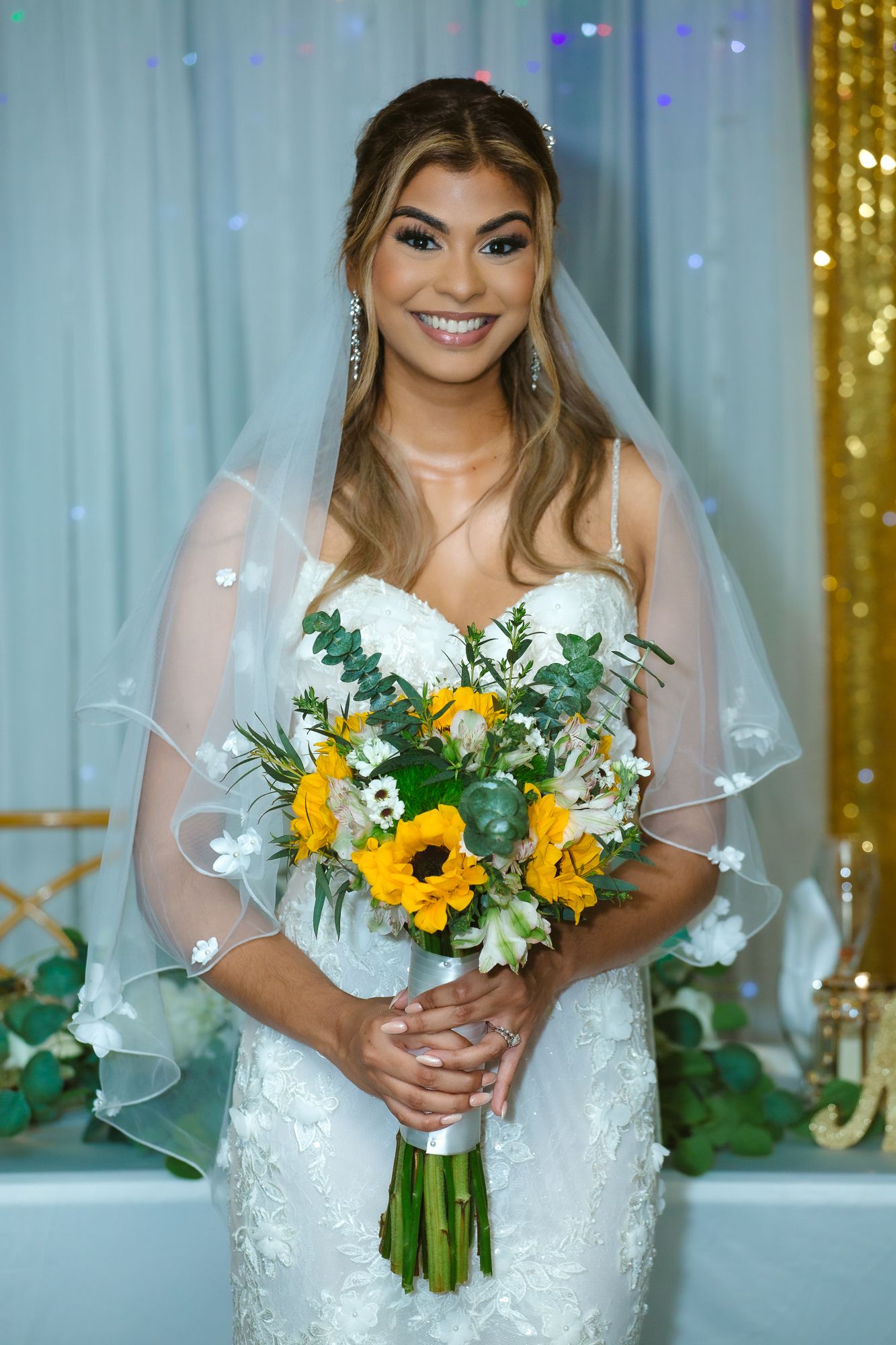 Radiant bride holds yellow sunflower bouquet while posing against sparkly gold backdrop.