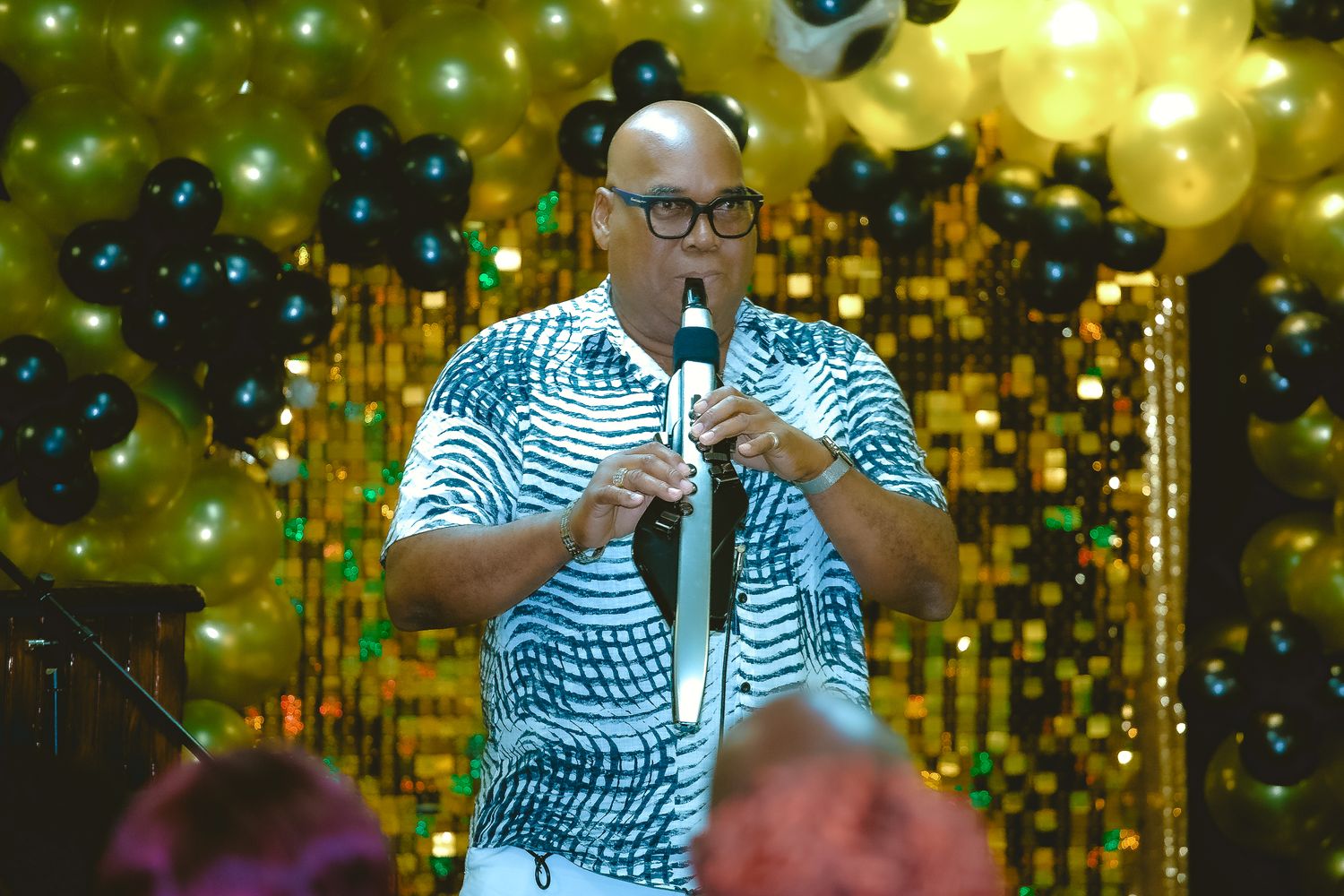 Performer in patterned shirt singing on stage with gold and black balloons in background.