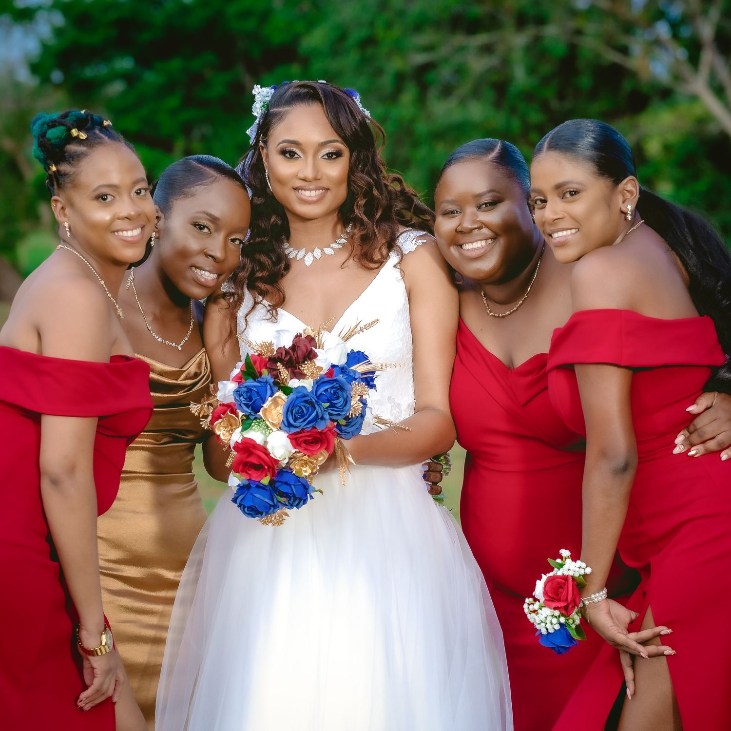 Bride in white gown poses with bridesmaids wearing red and gold dresses holding red and blue bouquets.