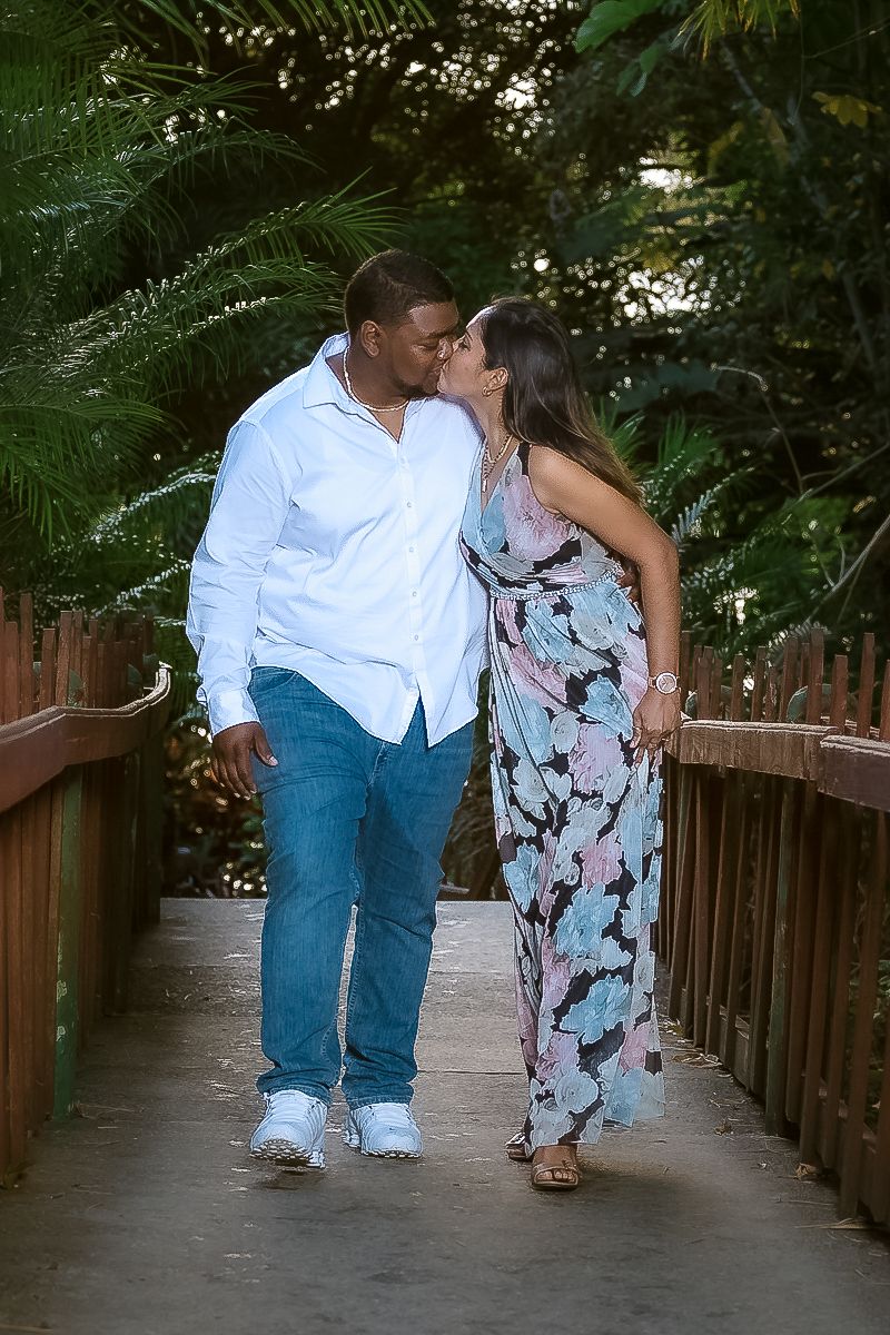 A romantic couple shares a tender moment while walking together on a wooden boardwalk surrounded by lush palm fronds.