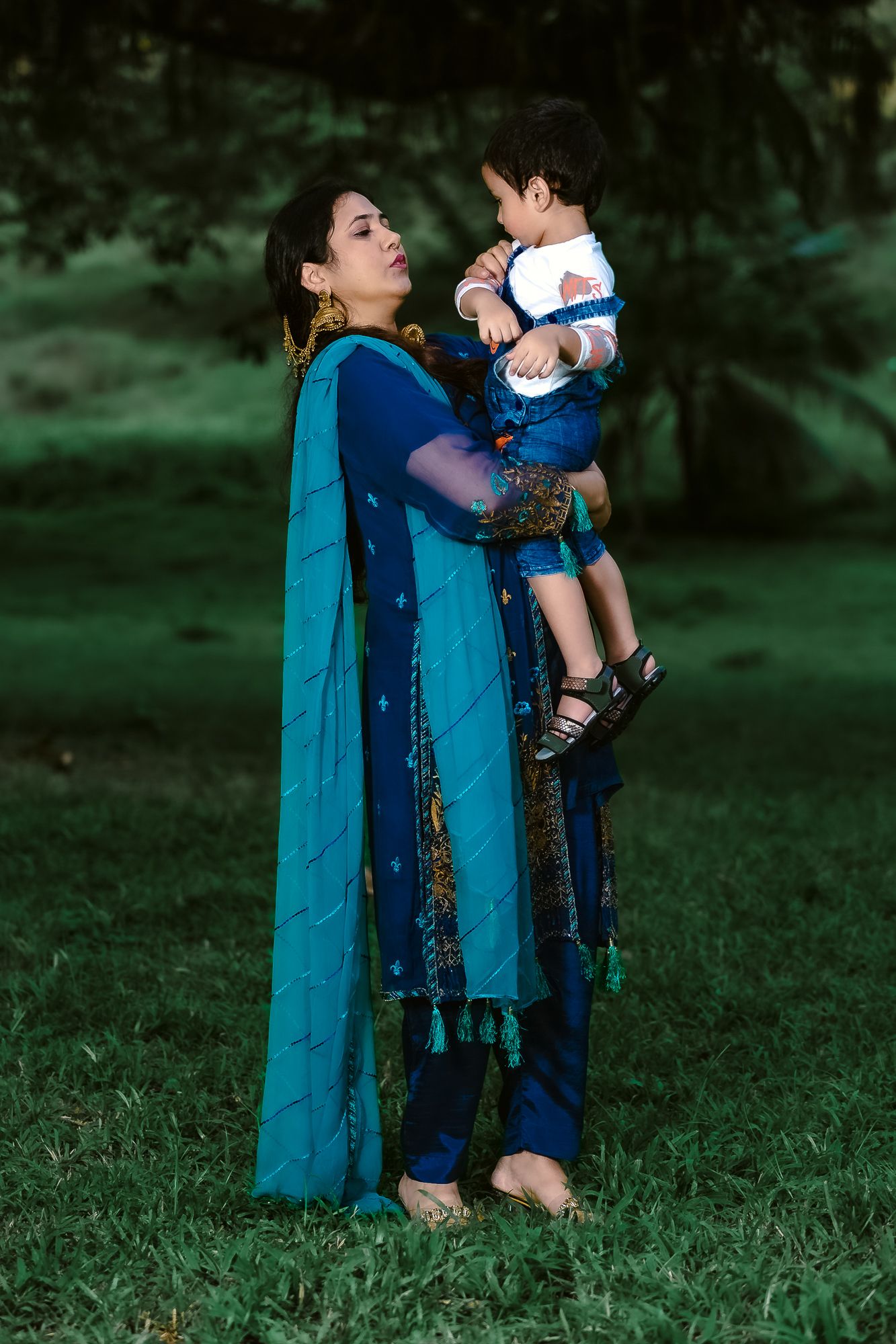 A woman in traditional blue attire holds a small child while standing in a grassy outdoor setting.