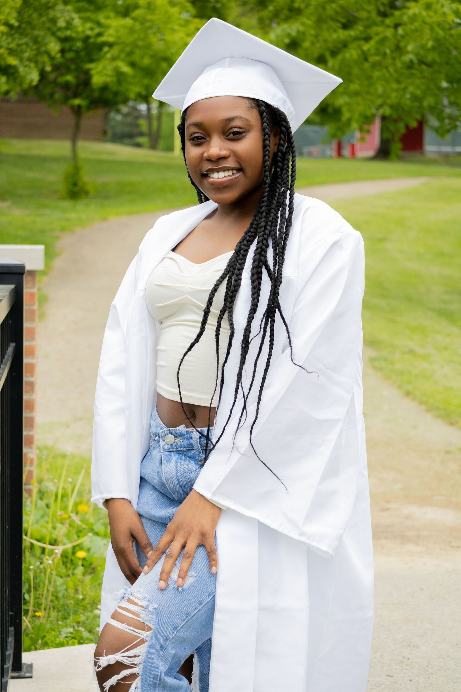 A graduate in white cap and gown and ripped jeans poses outdoors on a sunny day with trees in the background.