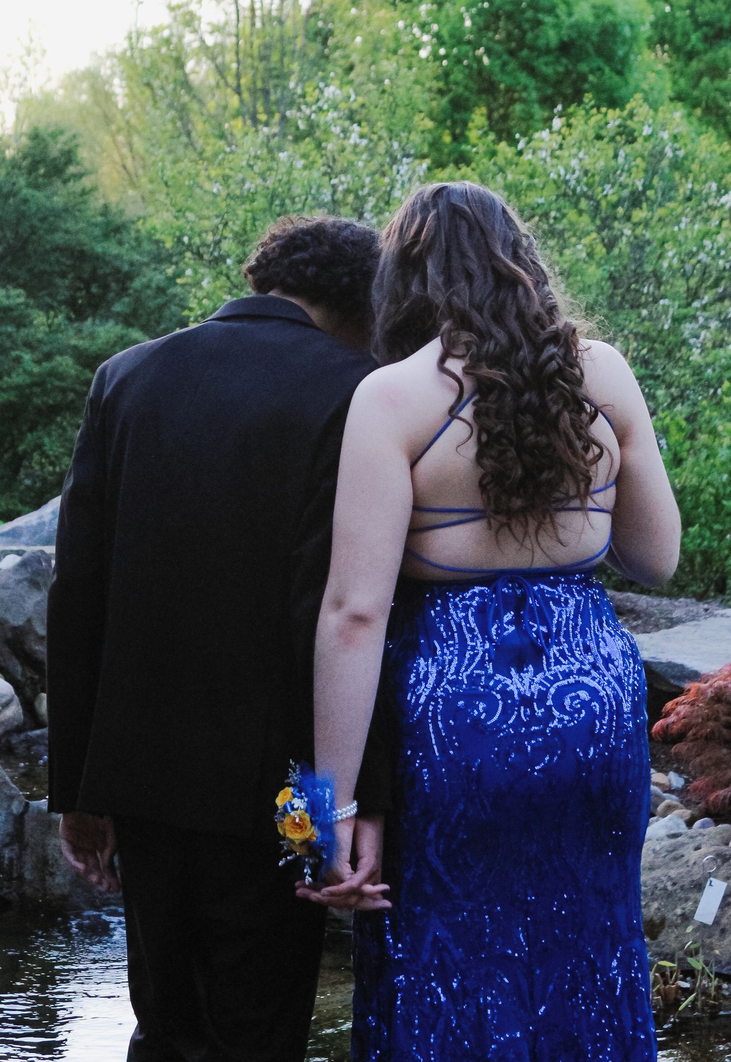 A couple in formal attire stands by a pond, with the person in a royal blue sequined gown holding a corsage.