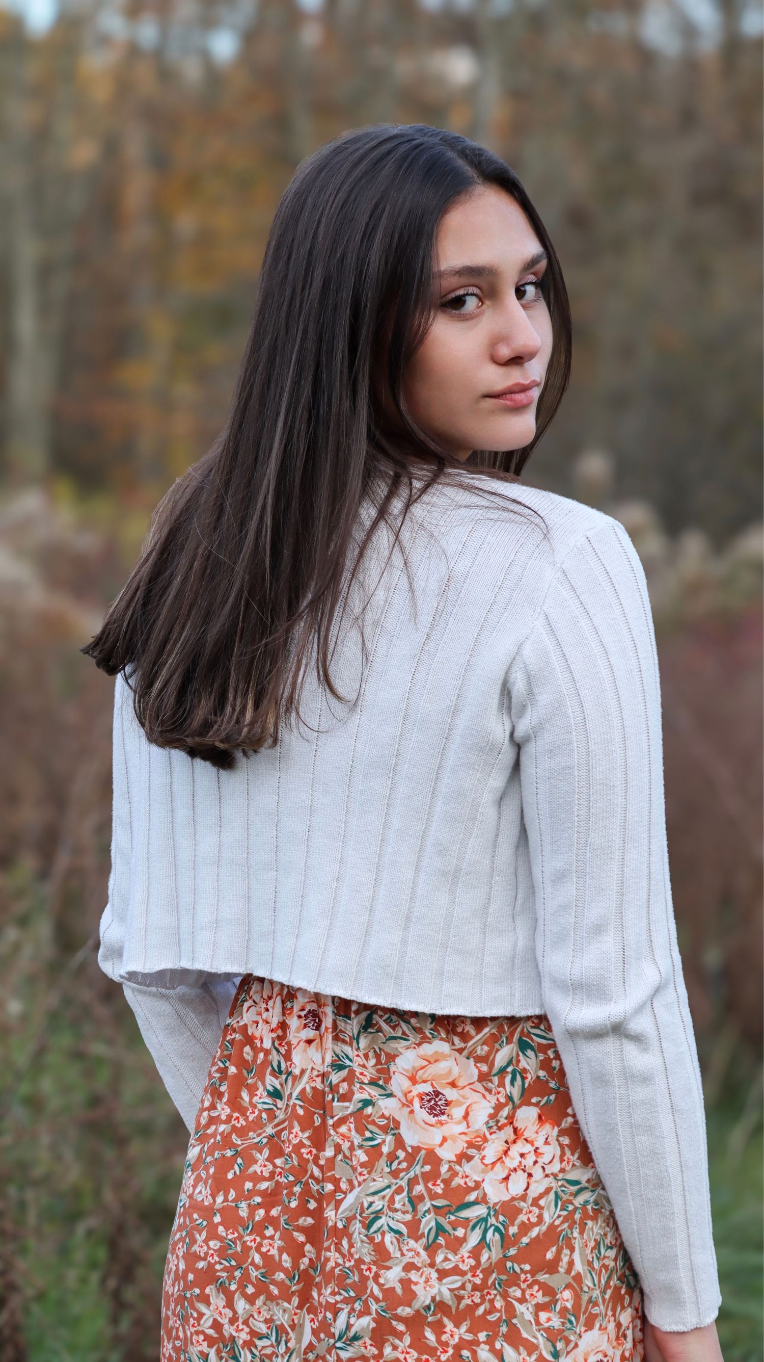 Someone in white sweater and floral dress poses against autumn foliage.