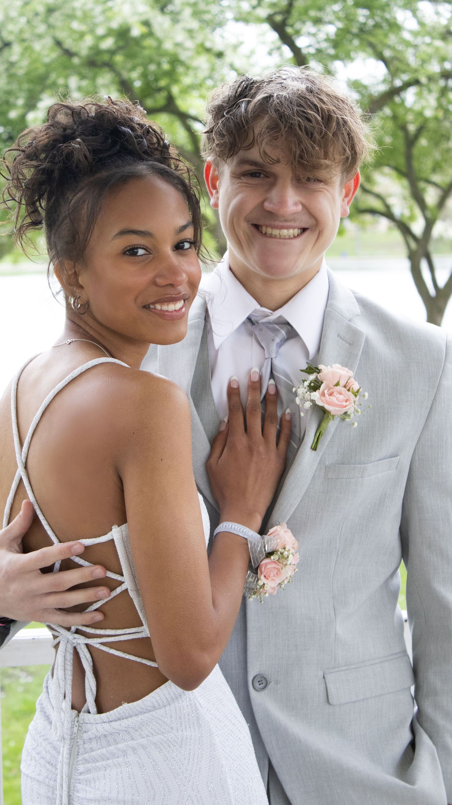 A couple in formal attire smiles together at what appears to be a prom or formal event outdoors.