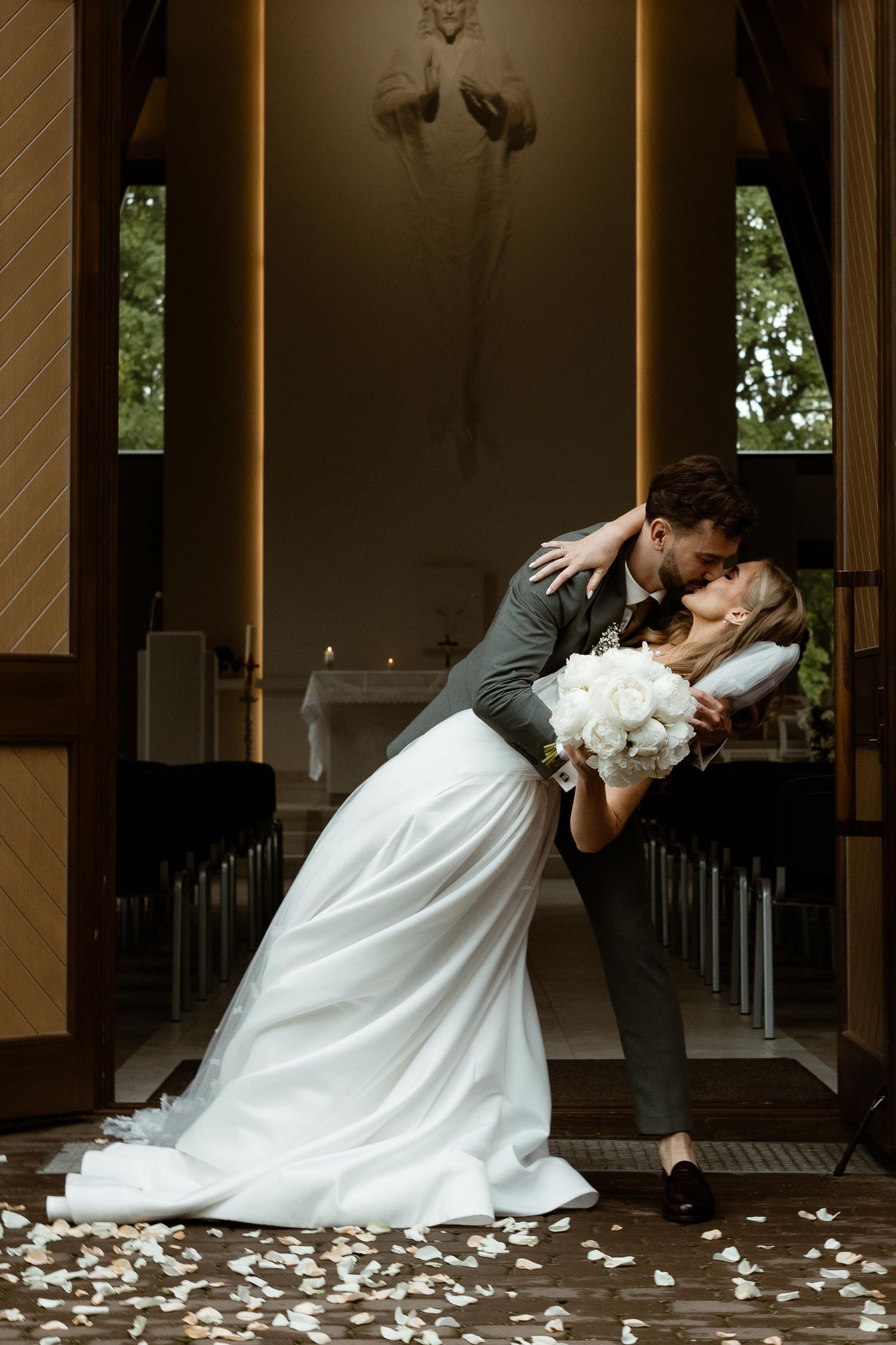 A newlywed couple shares a romantic dip kiss in a church aisle with white flower petals scattered on the ground.