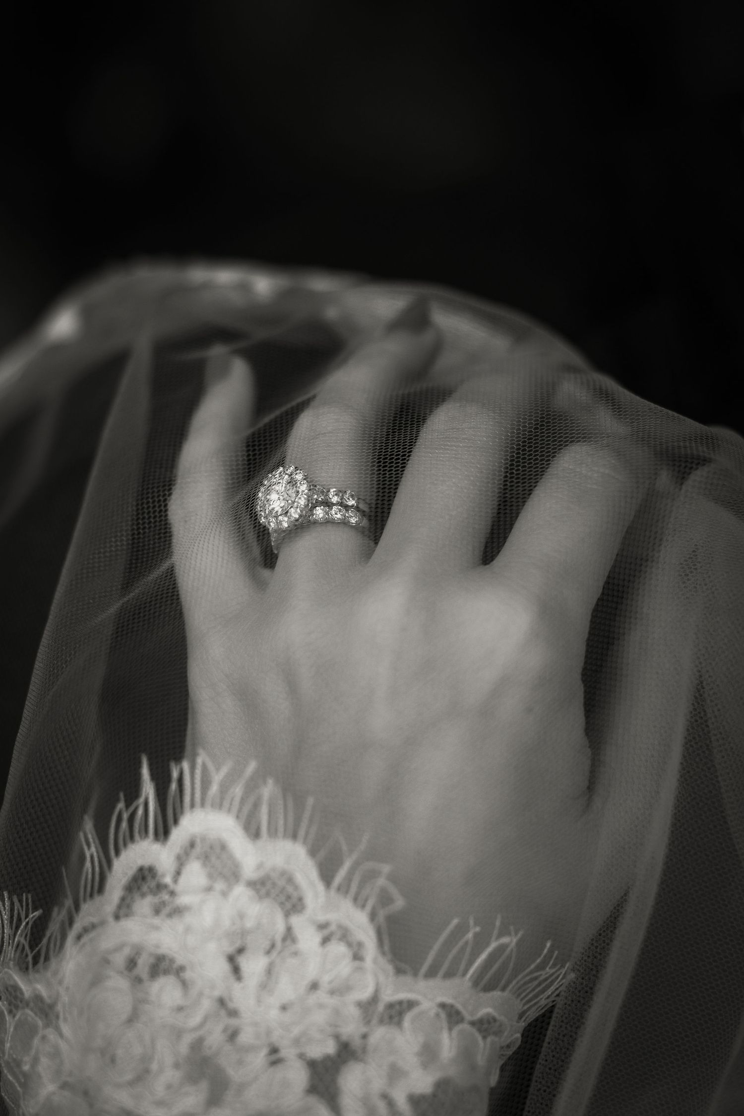 Black and white photo of a diamond engagement ring on a delicate hand resting against lace bridal veil.