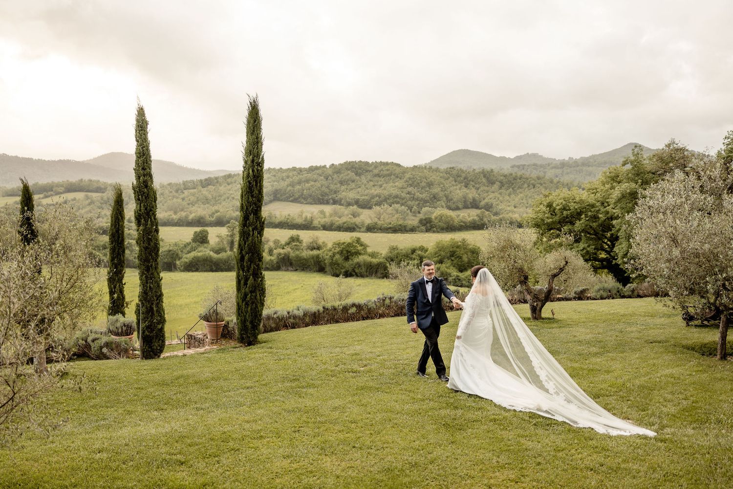 A bride in a flowing white dress walks down a scenic countryside path lined with cypress trees and rolling hills.