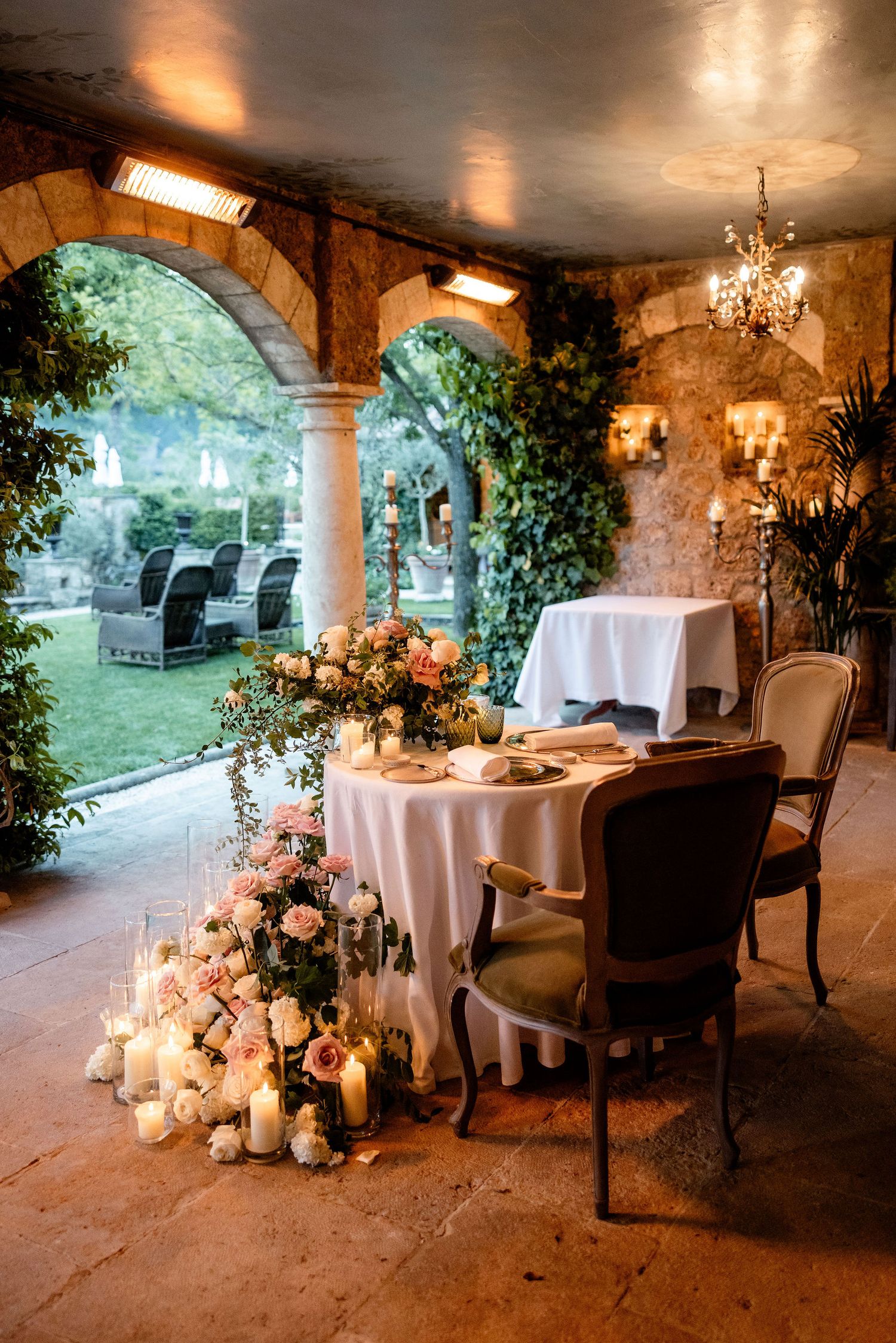 Romantic outdoor dining setup with candles and floral arrangements under stone arches at sunset.