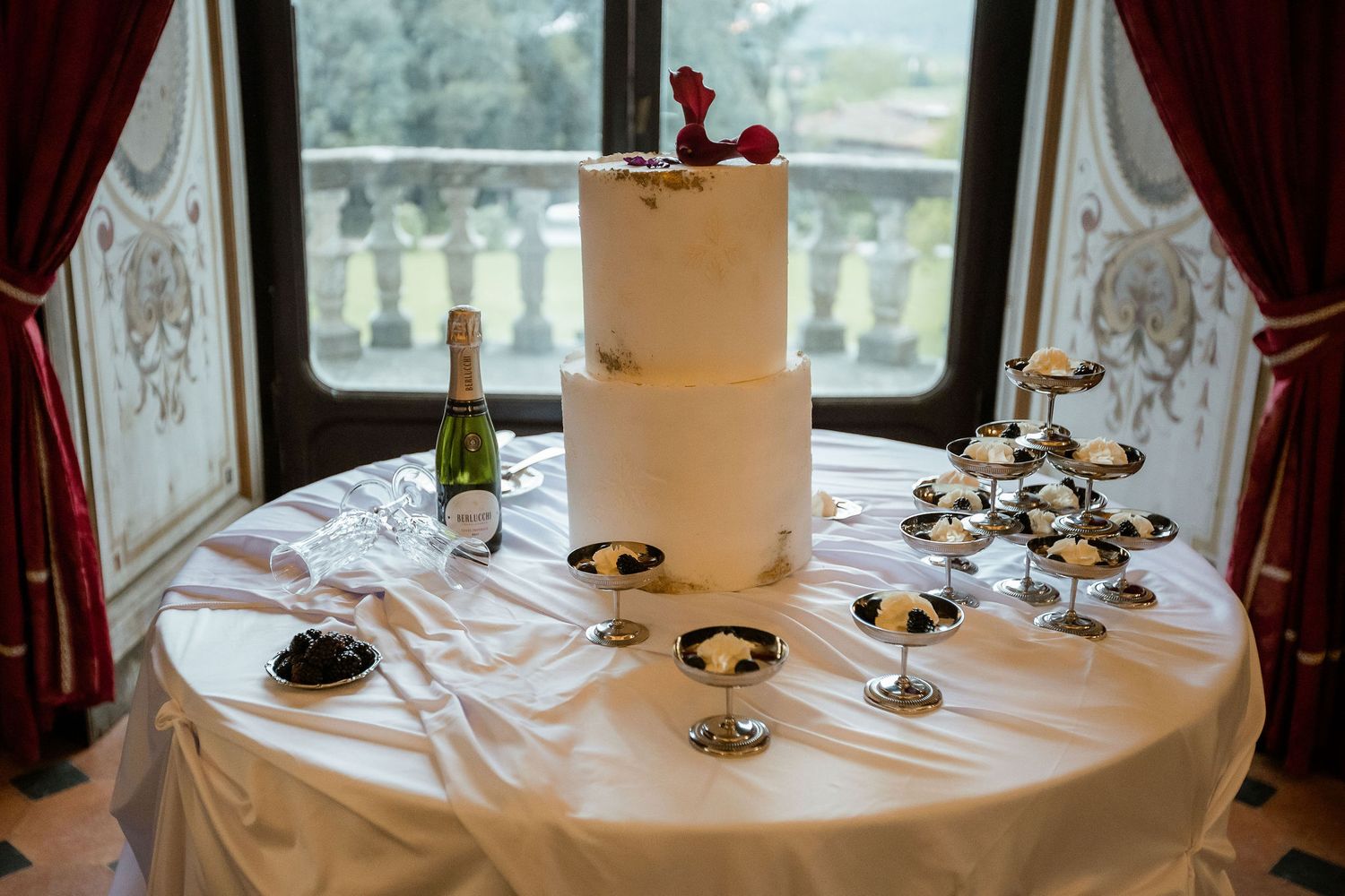 Elegant white wedding cake sits atop round table with dessert displays and red curtained window in background.