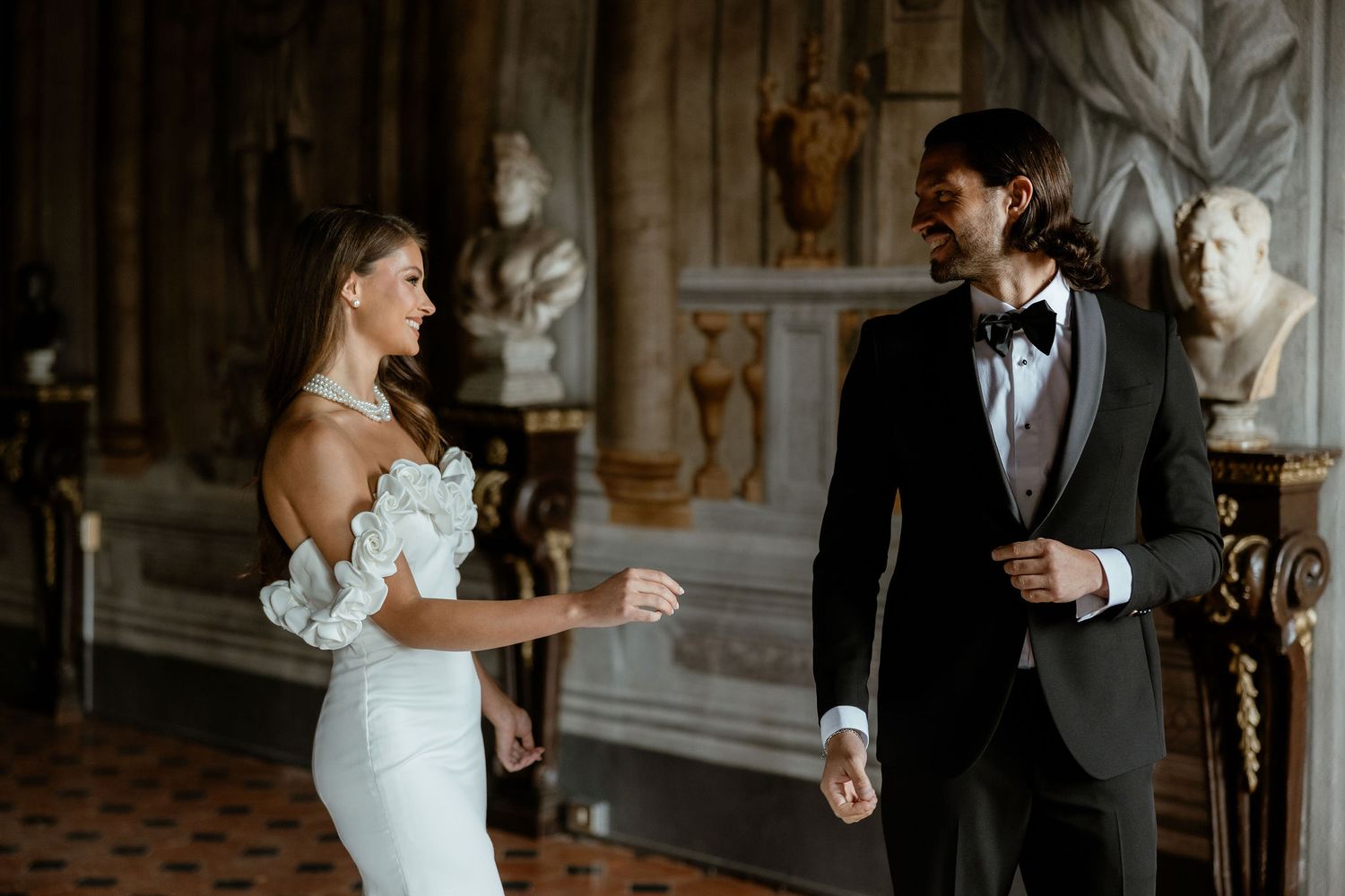 A bride and groom share their first look in an elegant room decorated with classical marble busts and ornate details.