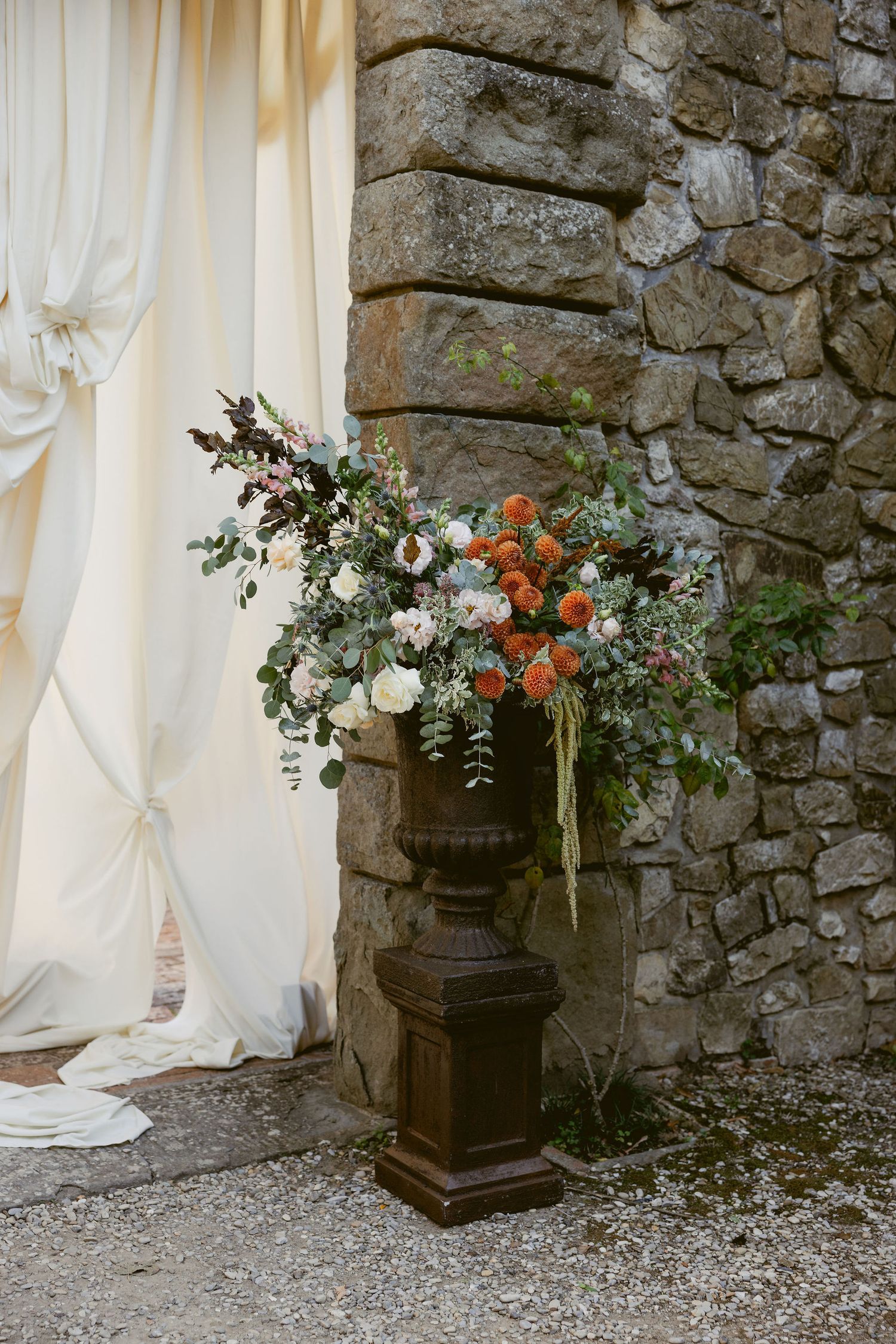Elegant floral arrangement with orange and white flowers in a dark pedestal urn against a stone wall and white curtain.