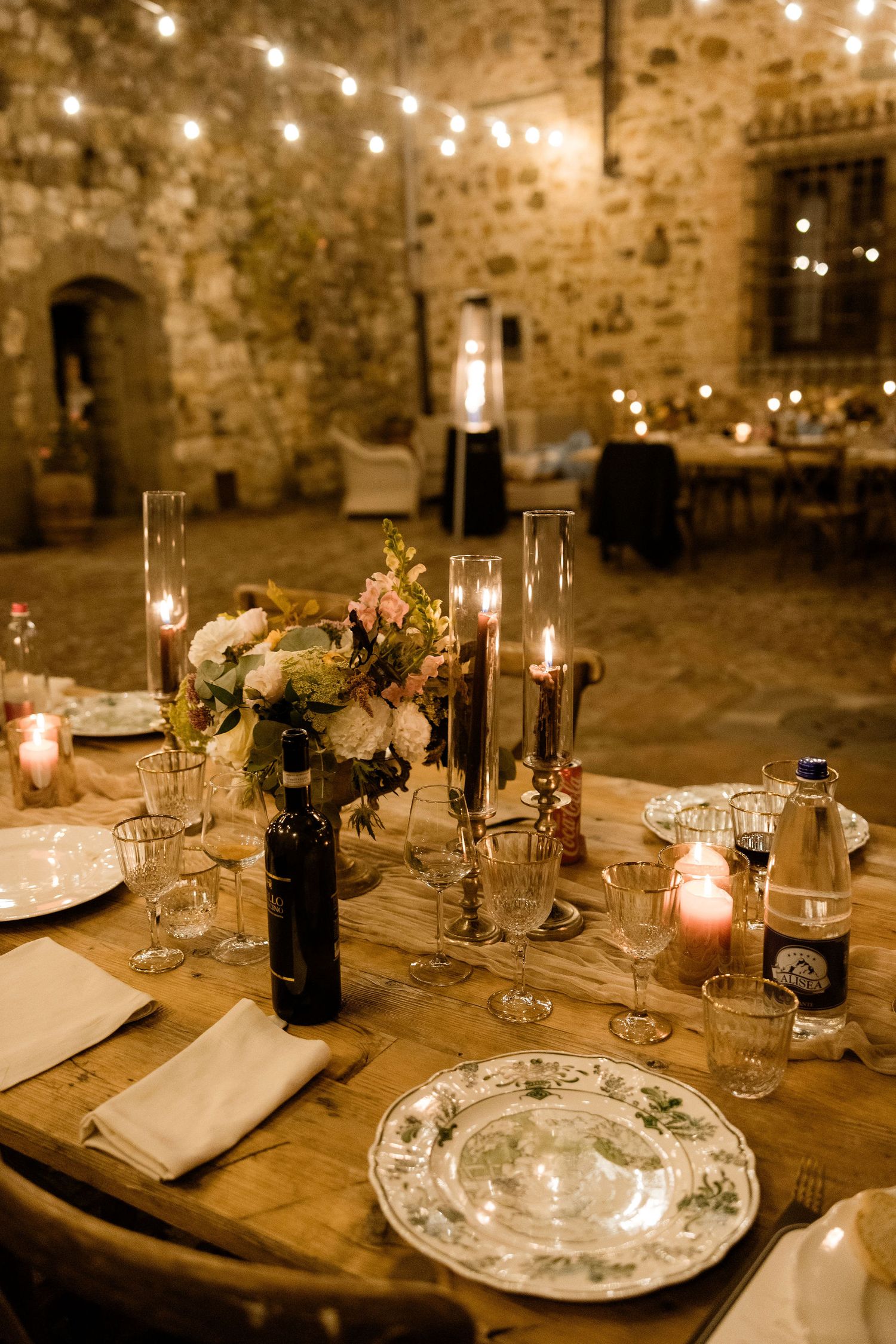 Rustic wedding table setting in stone castle with candlelight, wine bottles and floral centerpieces on wooden tables.