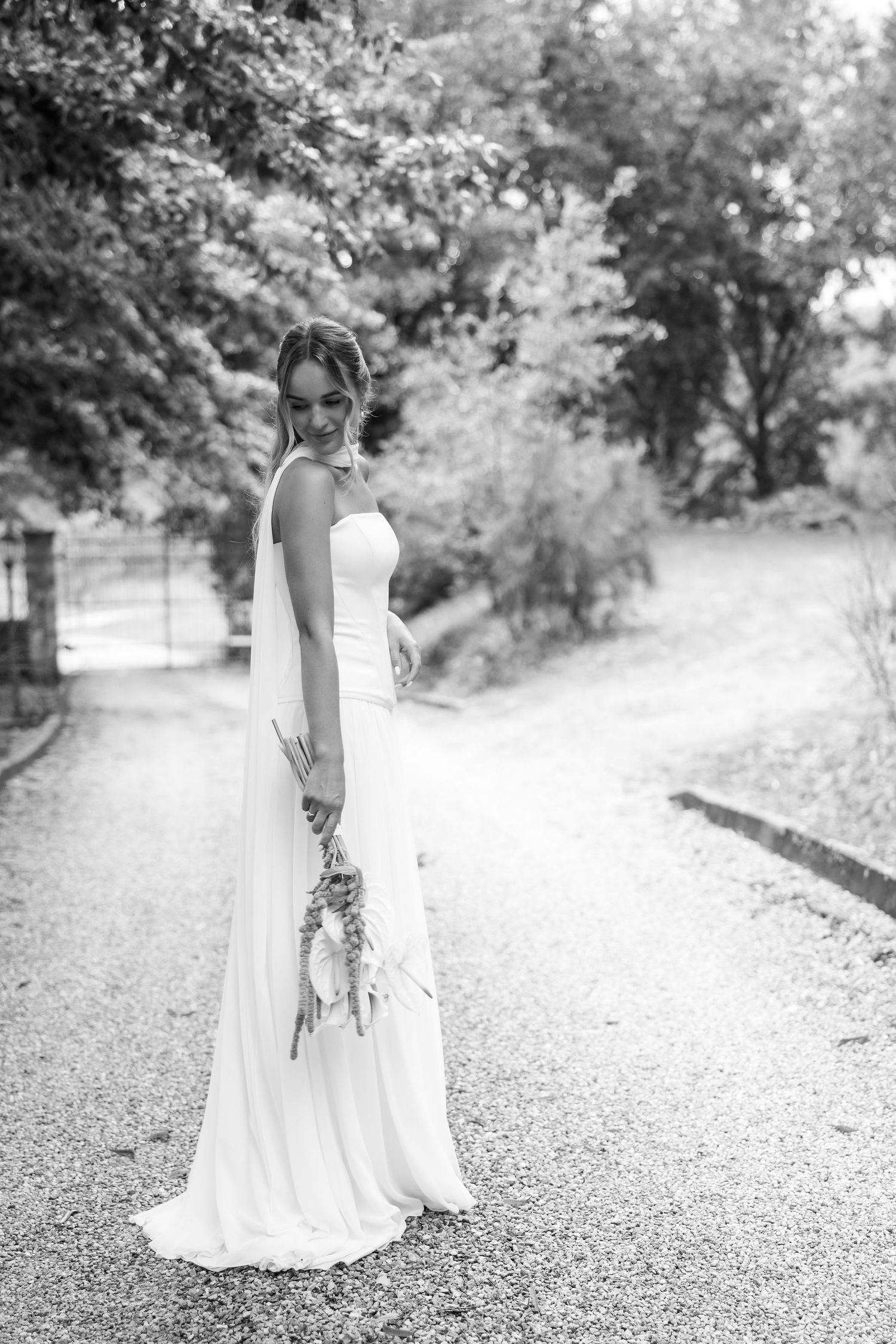 Black and white wedding photo of someone in a flowing white strapless gown holding a bouquet on a gravel path outdoors.