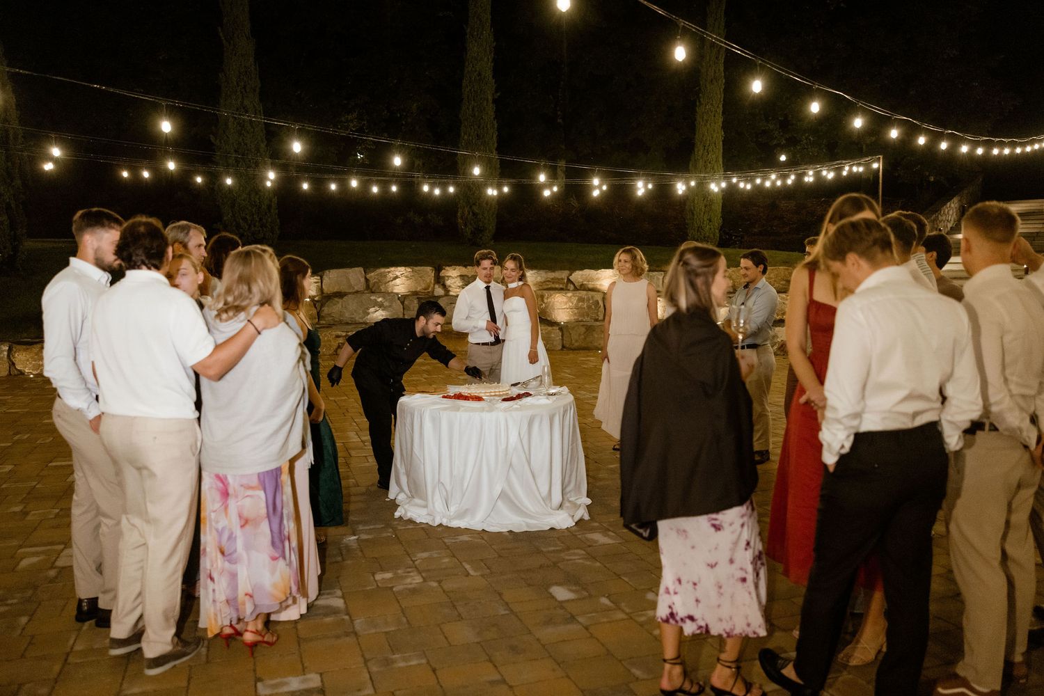 Wedding guests gather around a cake table under outdoor string lights at an evening reception celebration.