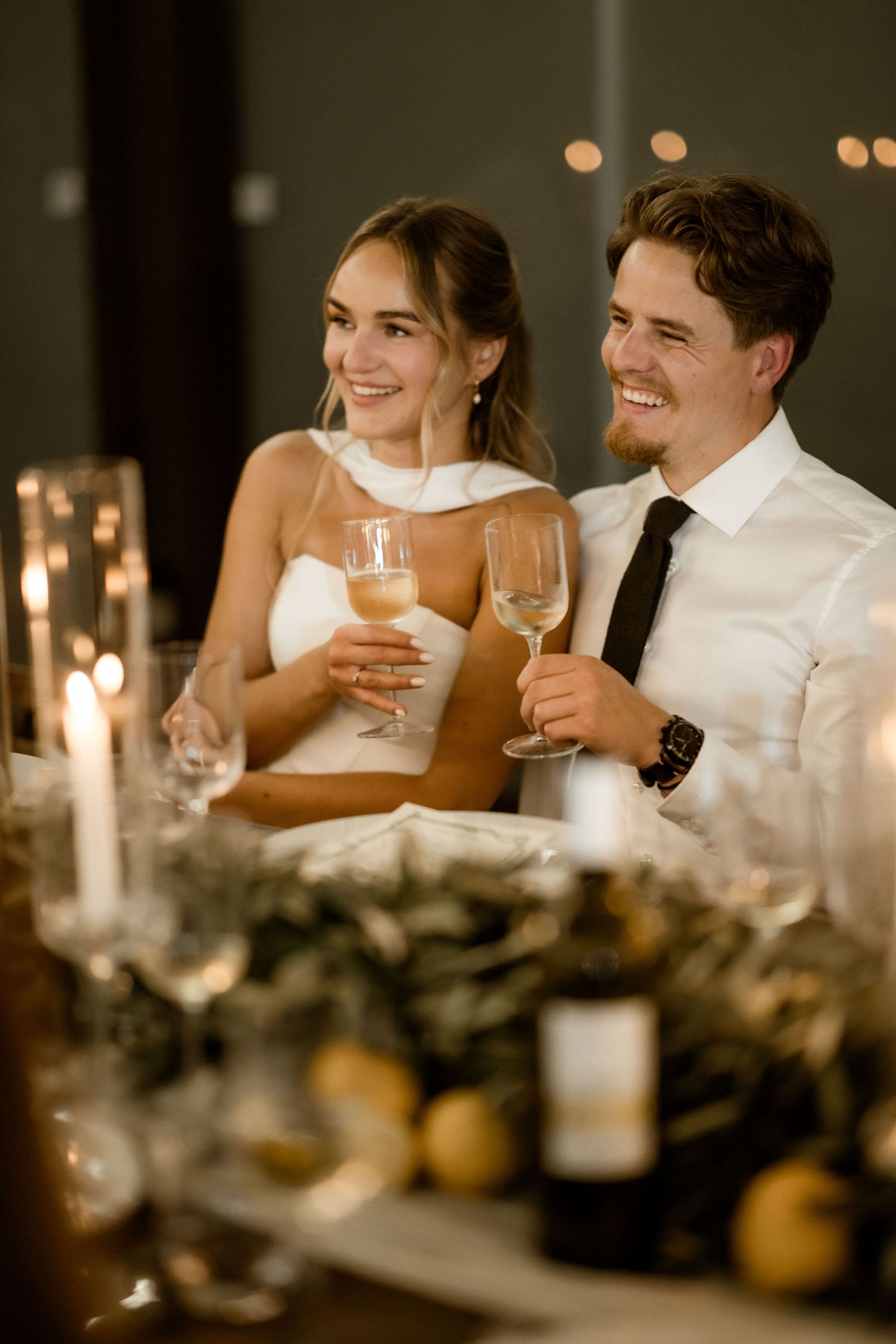 Wedding reception guests in white attire share joyful moments over drinks at an elegant candlelit dinner table.