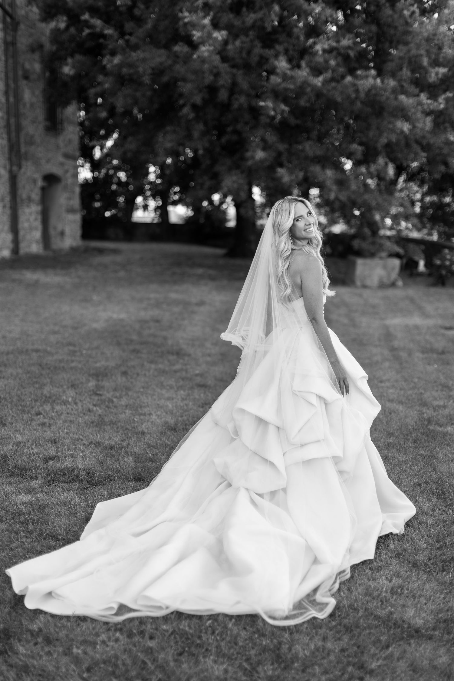 A flowing white wedding dress with a long train billows gracefully across a grassy lawn in black and white.