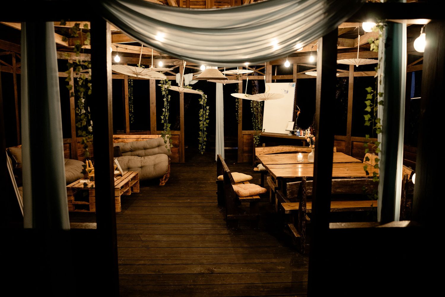 An intimate outdoor dining area with white draping, wooden deck flooring, and warm lighting under a covered pavilion.