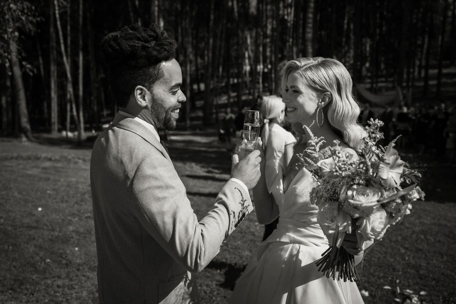 Black and white photo of intimate wedding moment in forest setting with floral bouquet.
